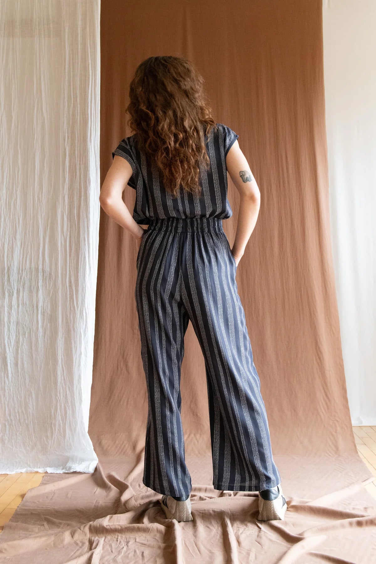 Back view of a woman wearing the Padua Pants by Kazak in Charcoal Dots, with a high waist, flat waistband at the front and elastic at the bag, wide legs, and large slanted pockets. She is wearing them with the matching Sandrigo top and standing in front of a brown curtain.