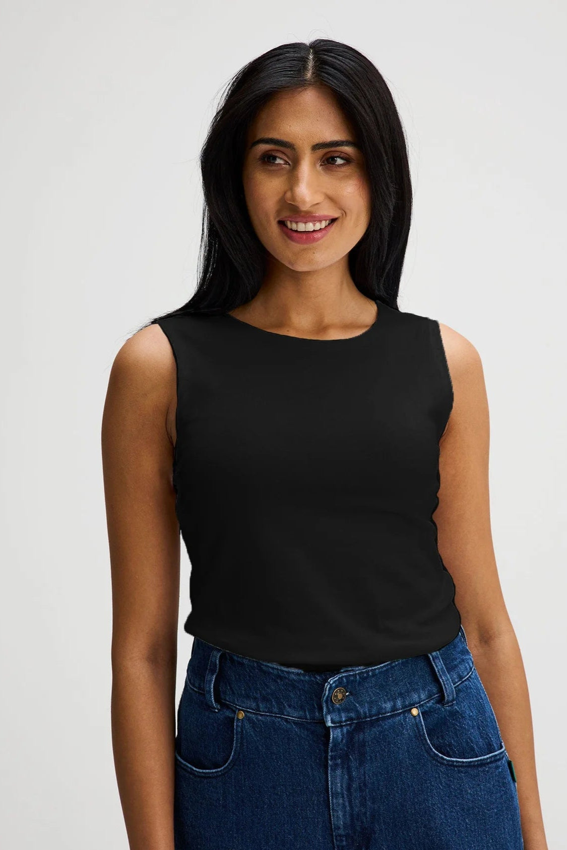 A woman wearing the Brompton Tank by Message Factory in Black, a crew neck tank with whide straps and a slim fit in organic cotton. She is wearing it with jeans and standing in front of a white background.