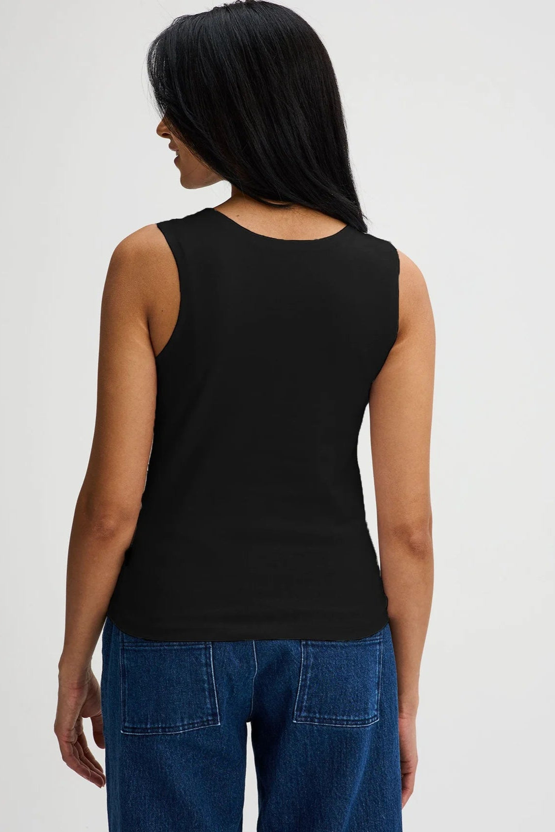 Back view of a woman wearing the Brompton Tank by Message Factory in Black, a crew neck tank with wide straps and a slim fit in organic cotton. She is wearing it with jeans and standing in front of a white background.