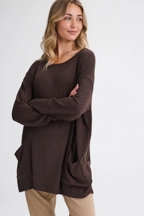 A woman wearing the Auclair Top by Message Factory in Brown, a bamboo/cotton/modal knit with a wide neck, long drop sleeves, a hi-low hem, and front patch pockets. She is wearing it with tan pants and standing in front of a white background.