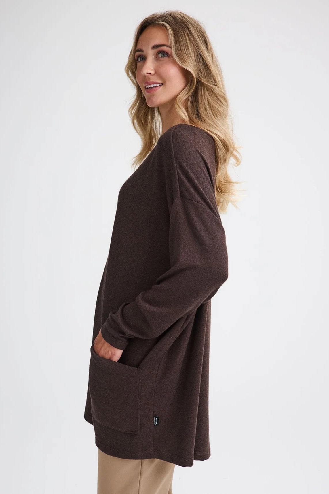 Side view of a woman wearing the Auclair Top by Message Factory in Brown, a bamboo/cotton/modal knit with a wide neck, long drop sleeves, a hi-low hem, and front patch pockets. She is wearing it with tan pants and standing in front of a white background.