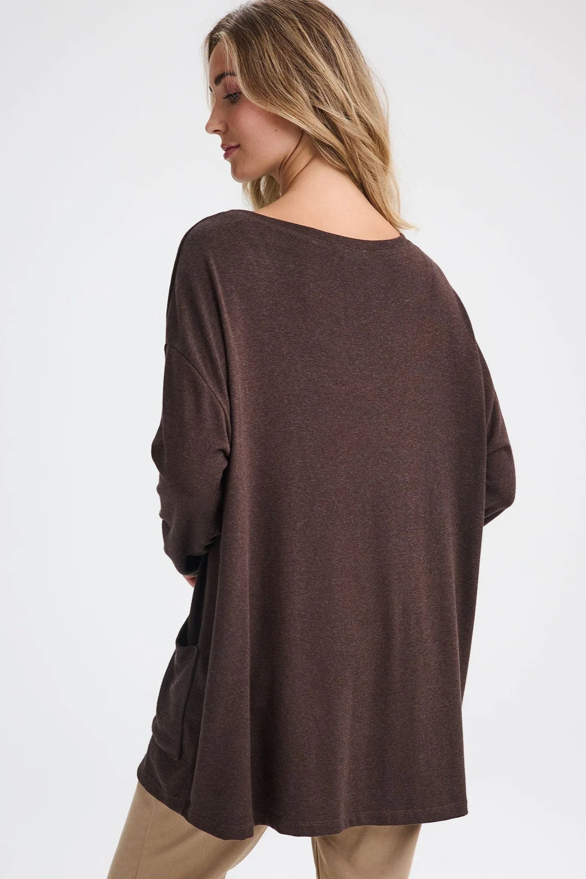 Back view of a woman wearing the Auclair Top by Message Factory in Brown, a bamboo/cotton/modal knit with a wide neck, long drop sleeves, a hi-low hem, and front patch pockets. She is wearing it with tan pants and standing in front of a white background.