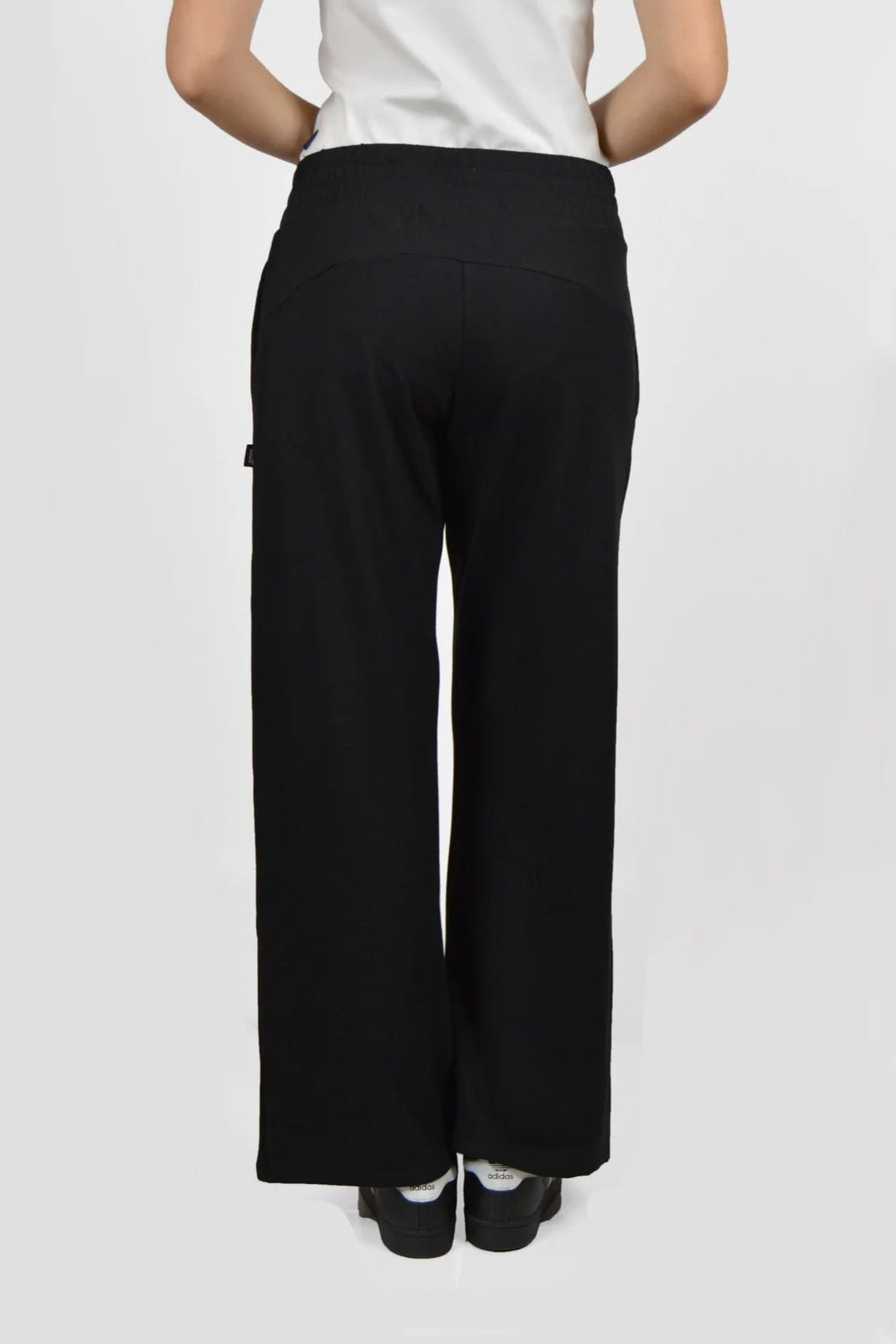 Back waist down view of the Tadoussac Pants by Message Factory in Black: flared sweatpants with an elastic waist in a bamboo/cotton blend. She is wearing a white tank with them and is standing in front of a white background.
