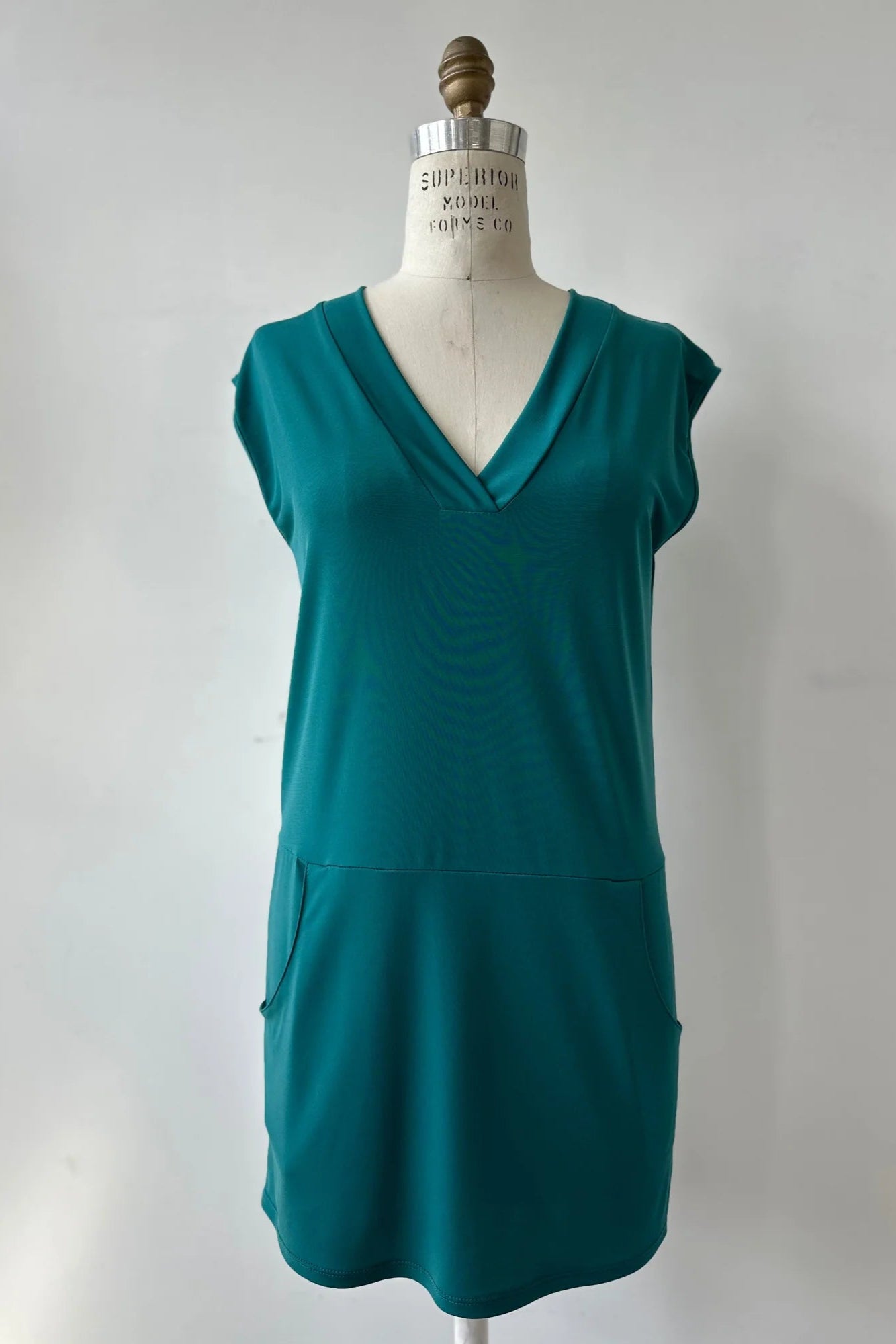 The Morticia Dress by Kollontai in Ocean, a sleeveless dress with a V-neck, a hi-low hemline, and a front pouch pocket, is show on a mannequin in front of a white background.