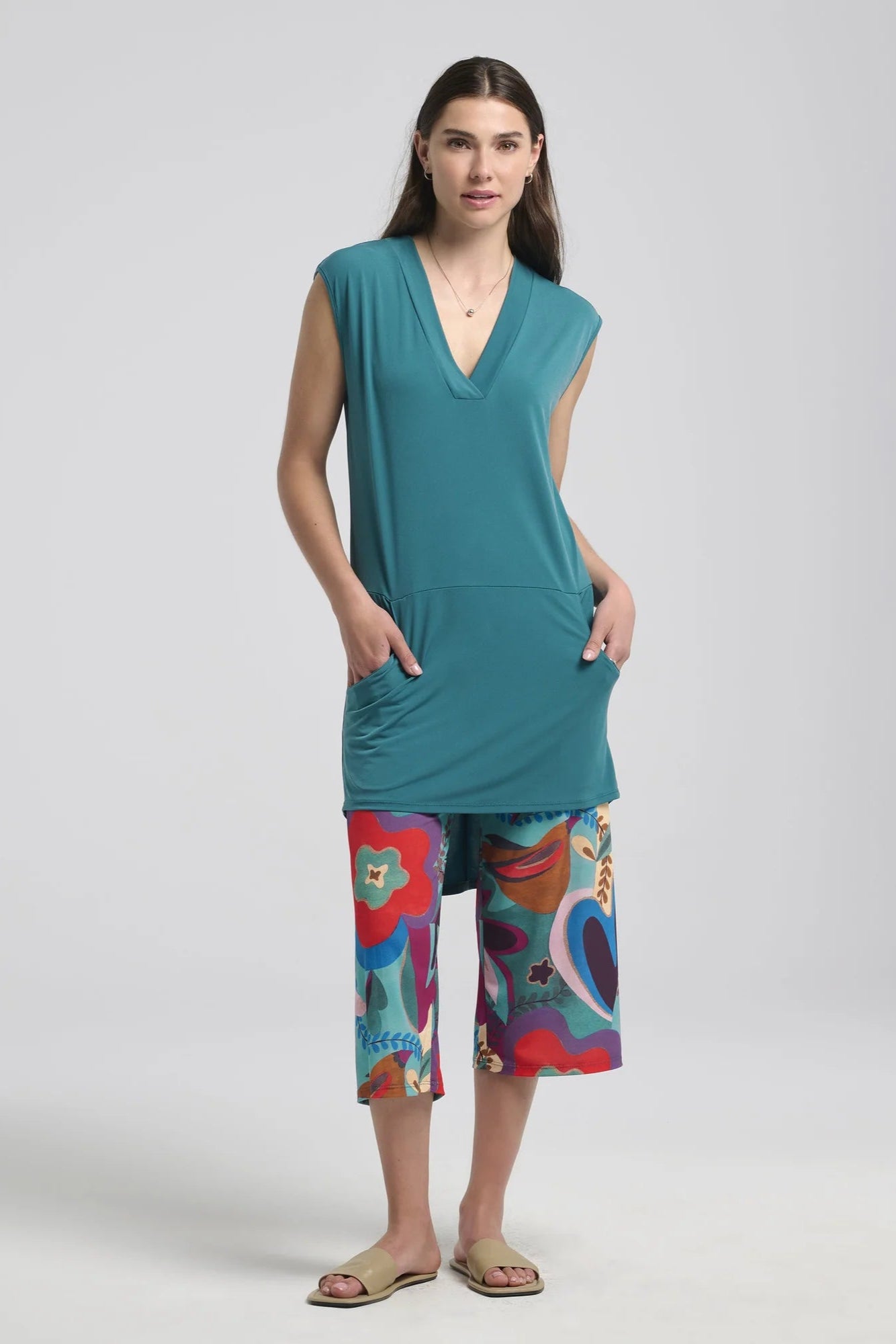A woman wearing the Morticia Dress by Kollontai in Ocean, a sleeveless dress with a V-neck, a hi-low hemline, and a front pouch pocket. She is wearing it with patterned cropped pants standing in front of a white background.