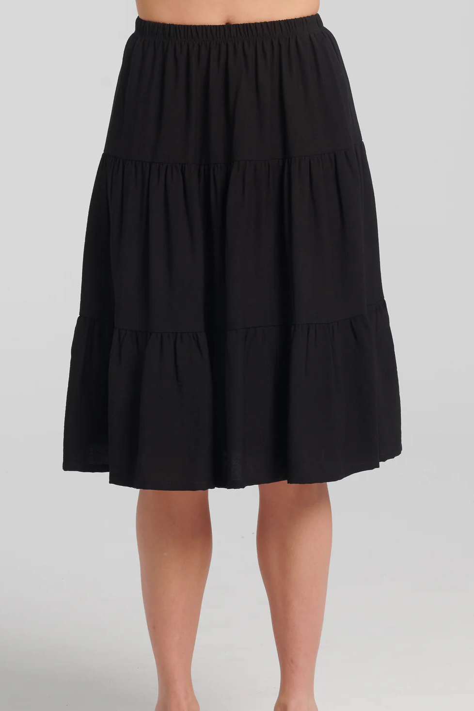 A waist-down view of a woman wearing the Al-Uzza Skirt by Kollontai in Black, a tiered skirt with an elastic waist and a below-the-knee length, standing in front of a white background