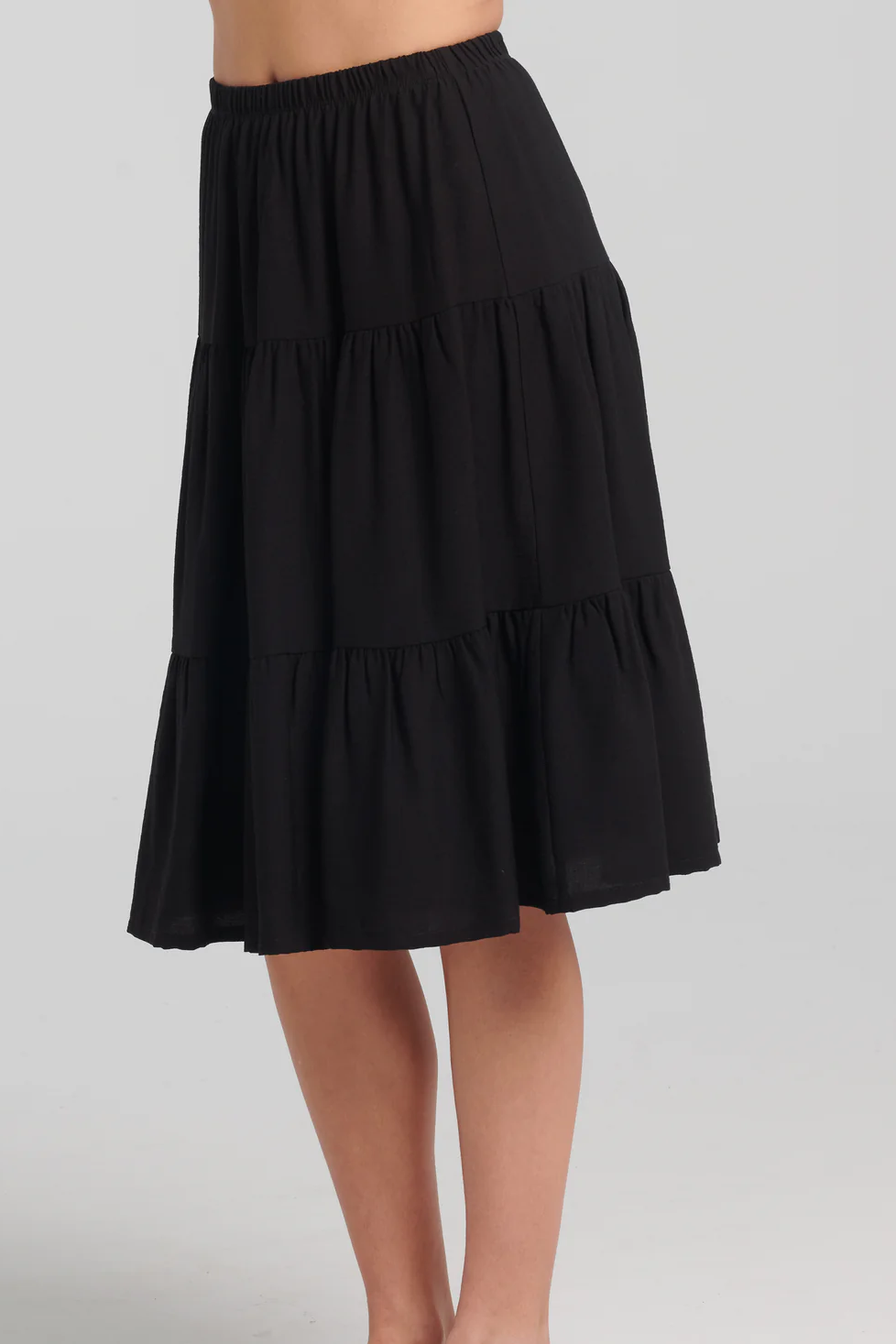 Waist down side view of a woman wearing the Al-Uzza Skirt by Kollontai in Black, a tiered skirt with an elastic waist and a below-the-knee length, standing against a white background