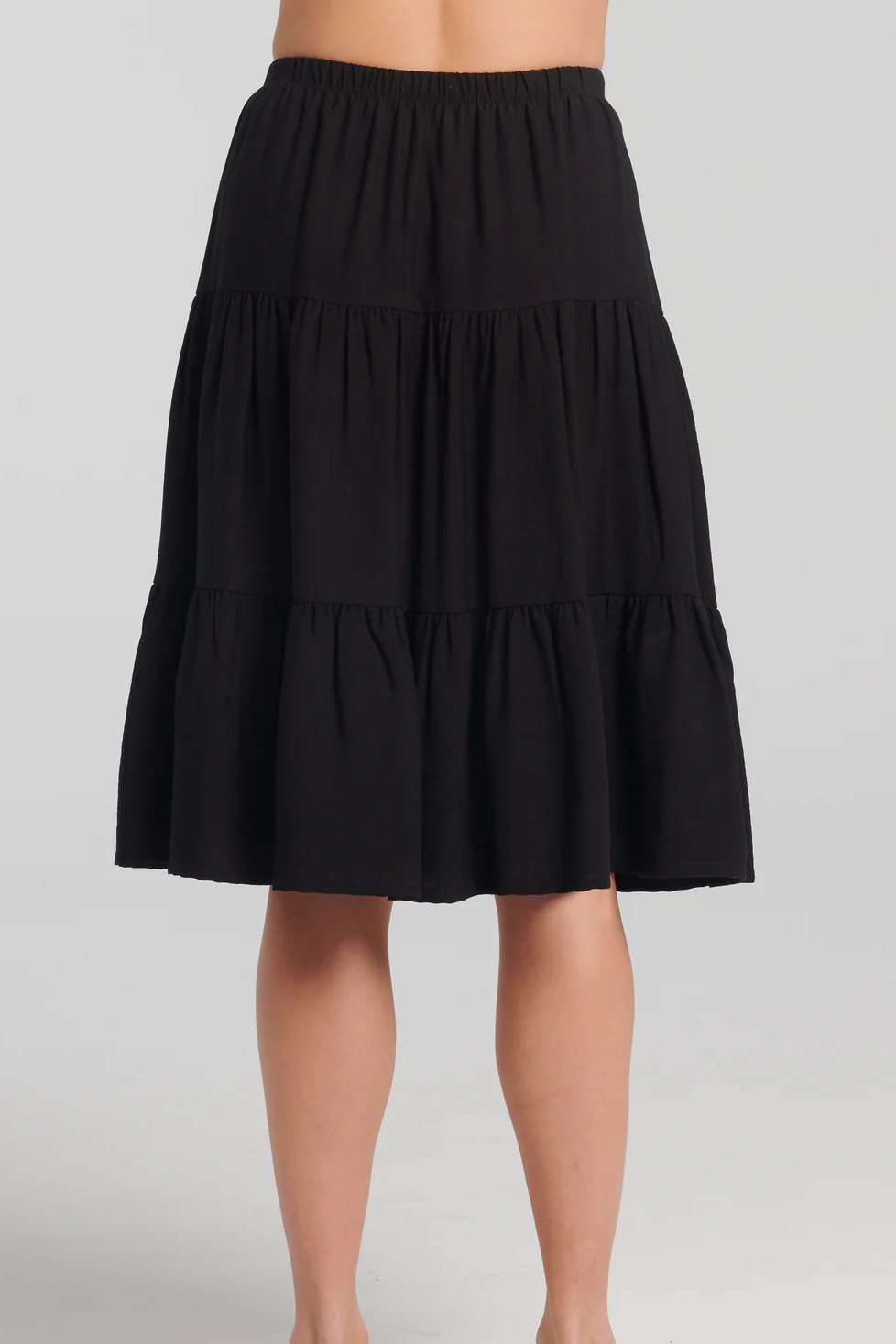 Back waist down view of a woman wearing the Al-Uzza Skirt by Kollontai in Black, a tiered skirt with an elastic waist and a below-the-knee length, standing in front of a white background