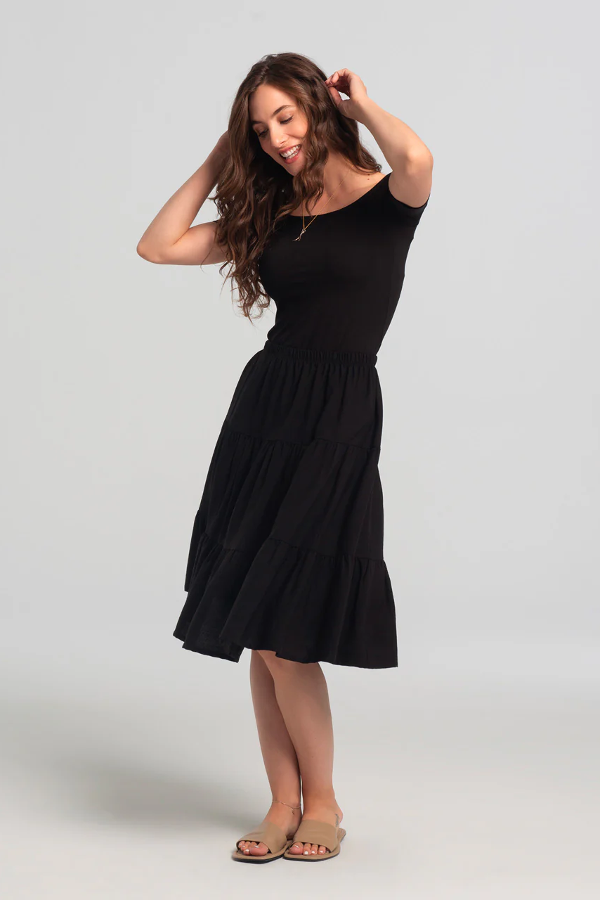 A woman wearing the Al-Uzza Skirt by Kollontai in Black, a tiered skirt with an elastic waist and a below-the-knee length, with a black top, standing in front of a white background
