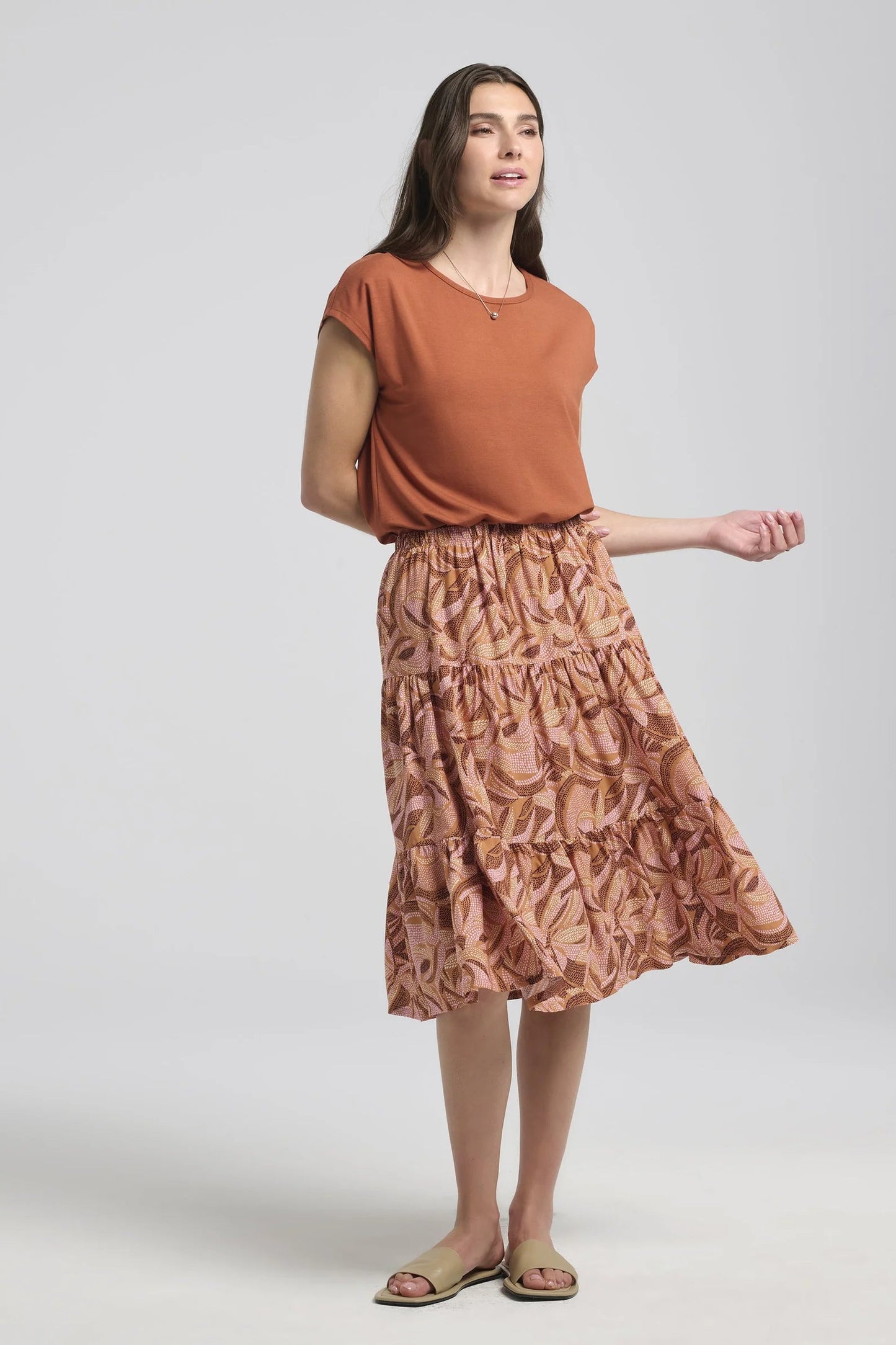 A woman wearing the Al-Uzza Skirt by Kollontai in Cinnamon print, a tiered skirt with an elastic waist and a below-the-knee length, with the Eowyn top in Rust. She is standing in front of a white background.