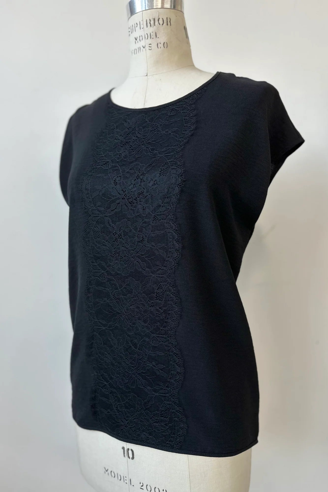 The Pearl T-shirt by Kollontai in Black, with short extended sleeves, a relaxed fit, and a lace applique down the front, is show on a mannequin in front of a white background.
