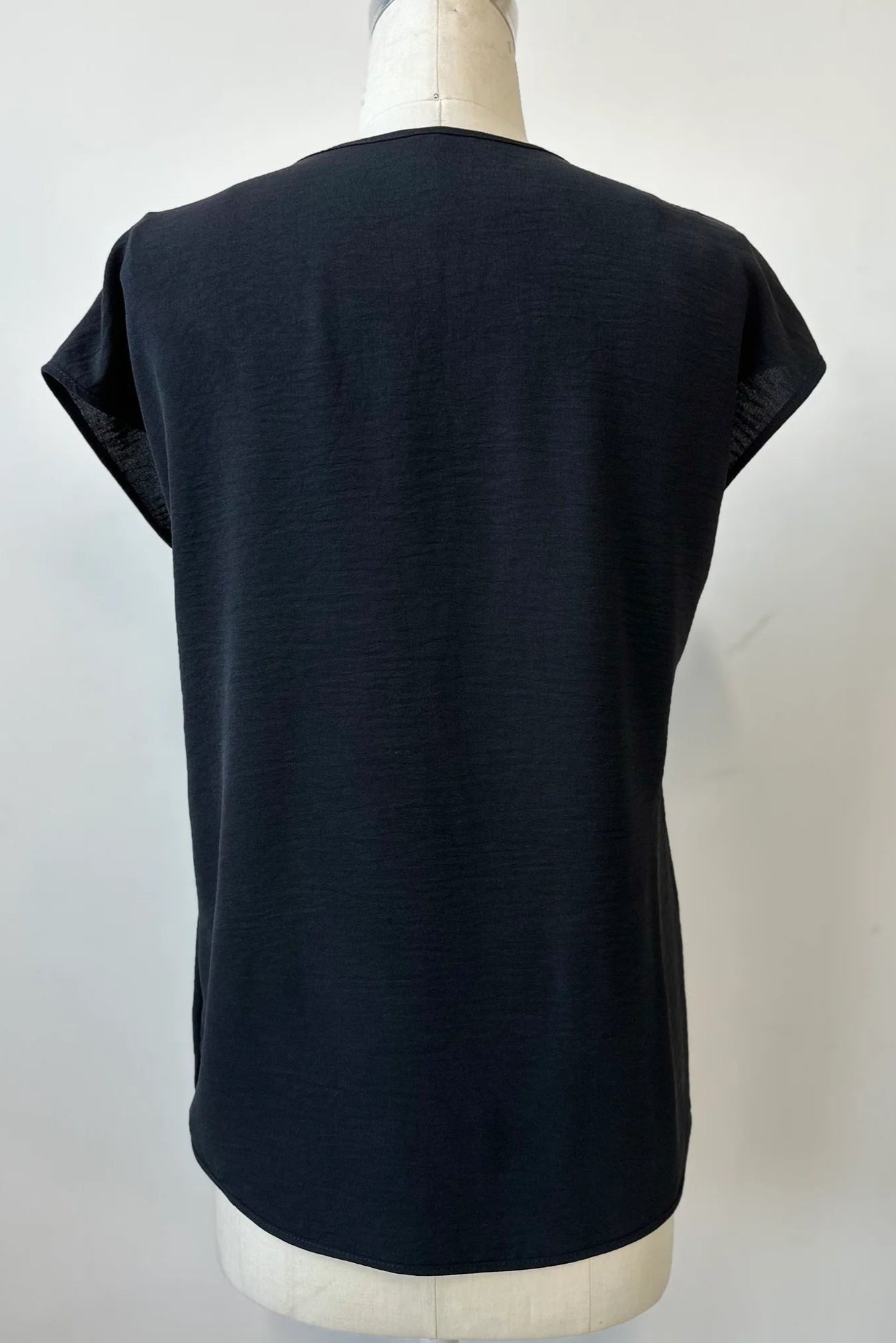 A back view of the Pearl T-shirt by Kollontai in Black, with short extended sleeves, a relaxed fit, and a lace applique down the front, is show on a mannequin in front of a white background.