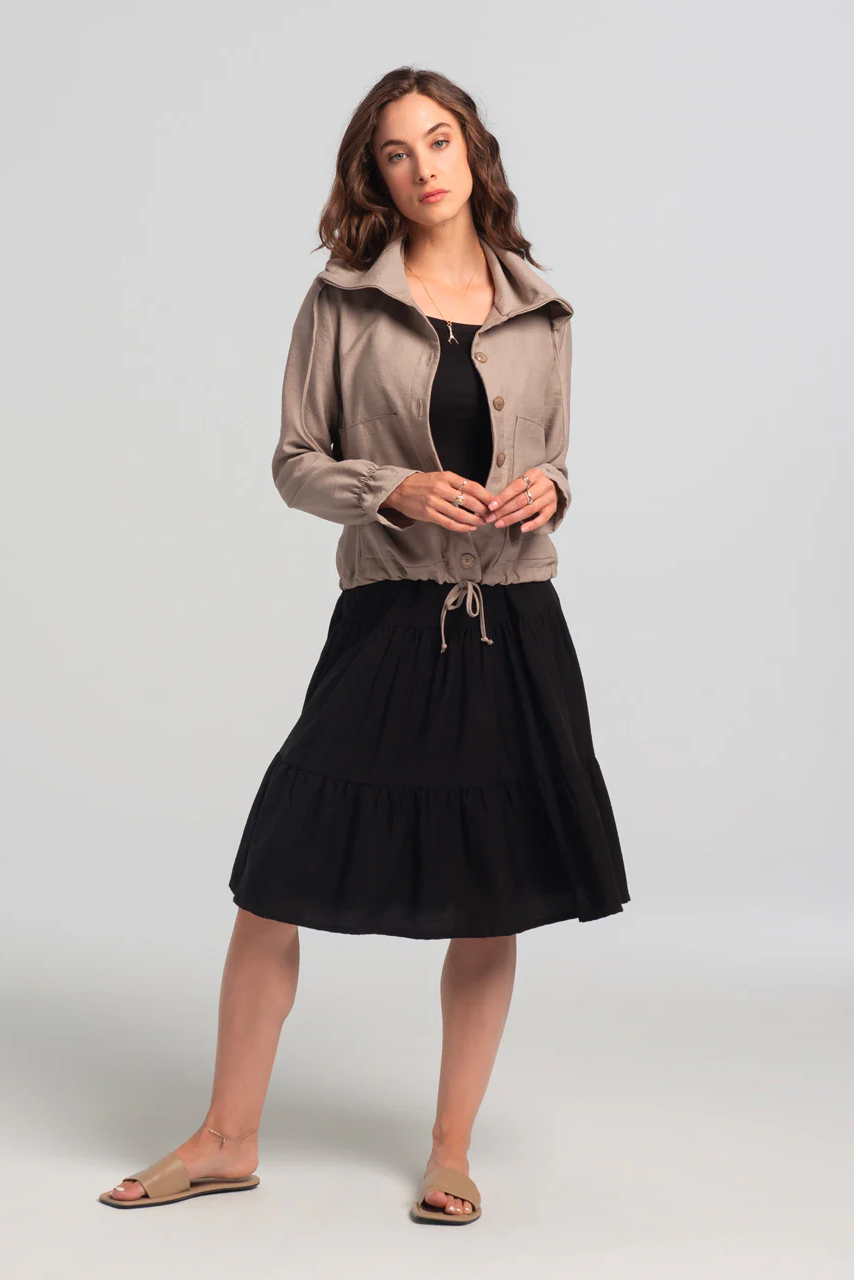 A woman wearing the Al-Uzza Skirt by Kollontai in Black, a tiered skirt with an elastic waist and a below-the-knee length, with a beige jacket, standing in front of a white background
