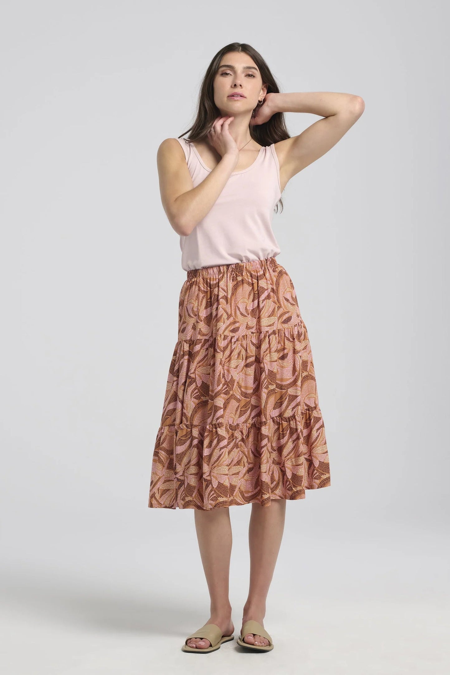 A woman wearing the Al-Uzza Skirt by Kollontai in Cinnamon print, a tiered skirt with an elastic waist and a below-the-knee length, with a pink top, standing in front of a white background