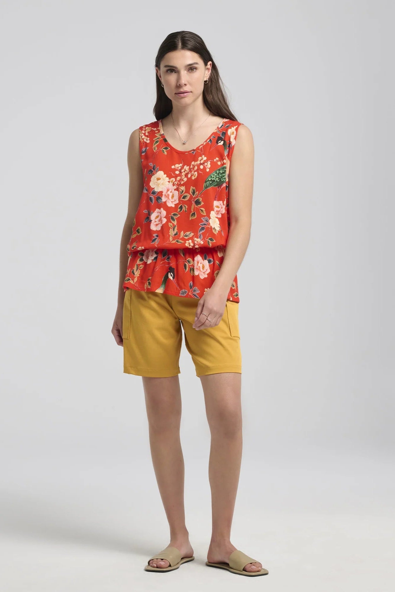 A woman wearing the Yvaine Top by Kollontai in Red, a sleeveless top with a scoop neck, wide straps, an elastic waist that creates a bloused effect. She is wearing it with the yellow shorts and is standing in front of a white background.