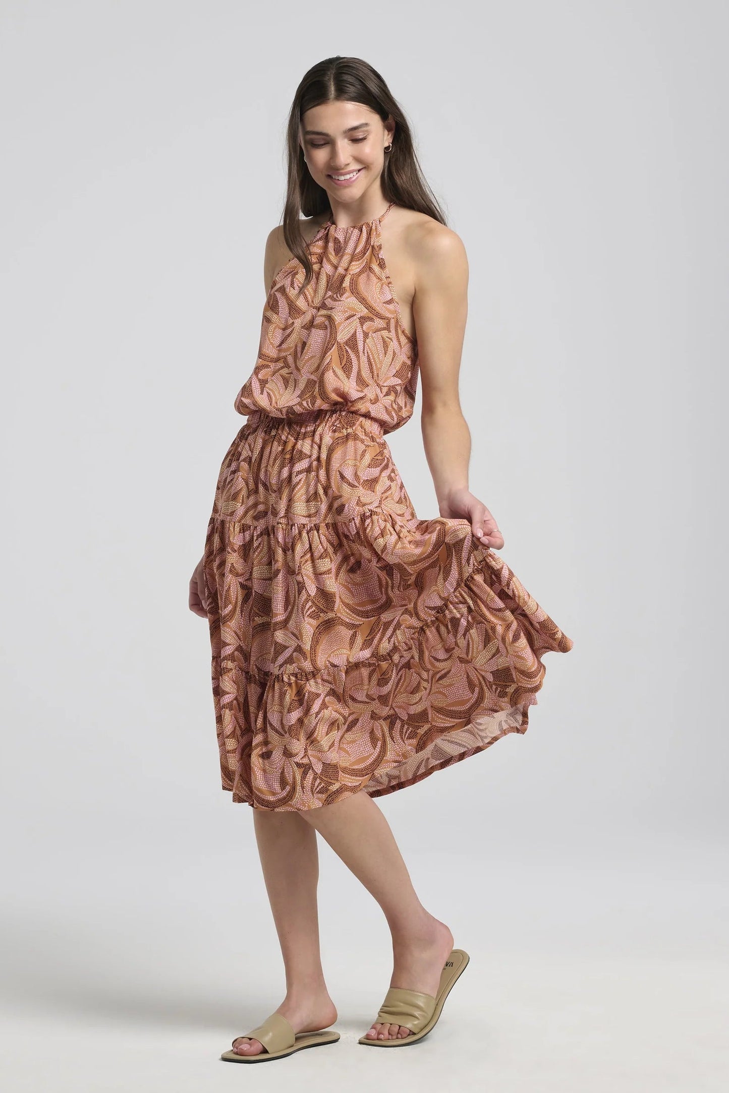 A woman wearing the Al-Uzza Skirt by Kollontai in Cinnamon print, a tiered skirt with an elastic waist and a below-the-knee length, with the matching Semira Camisole, standing in front of a white background