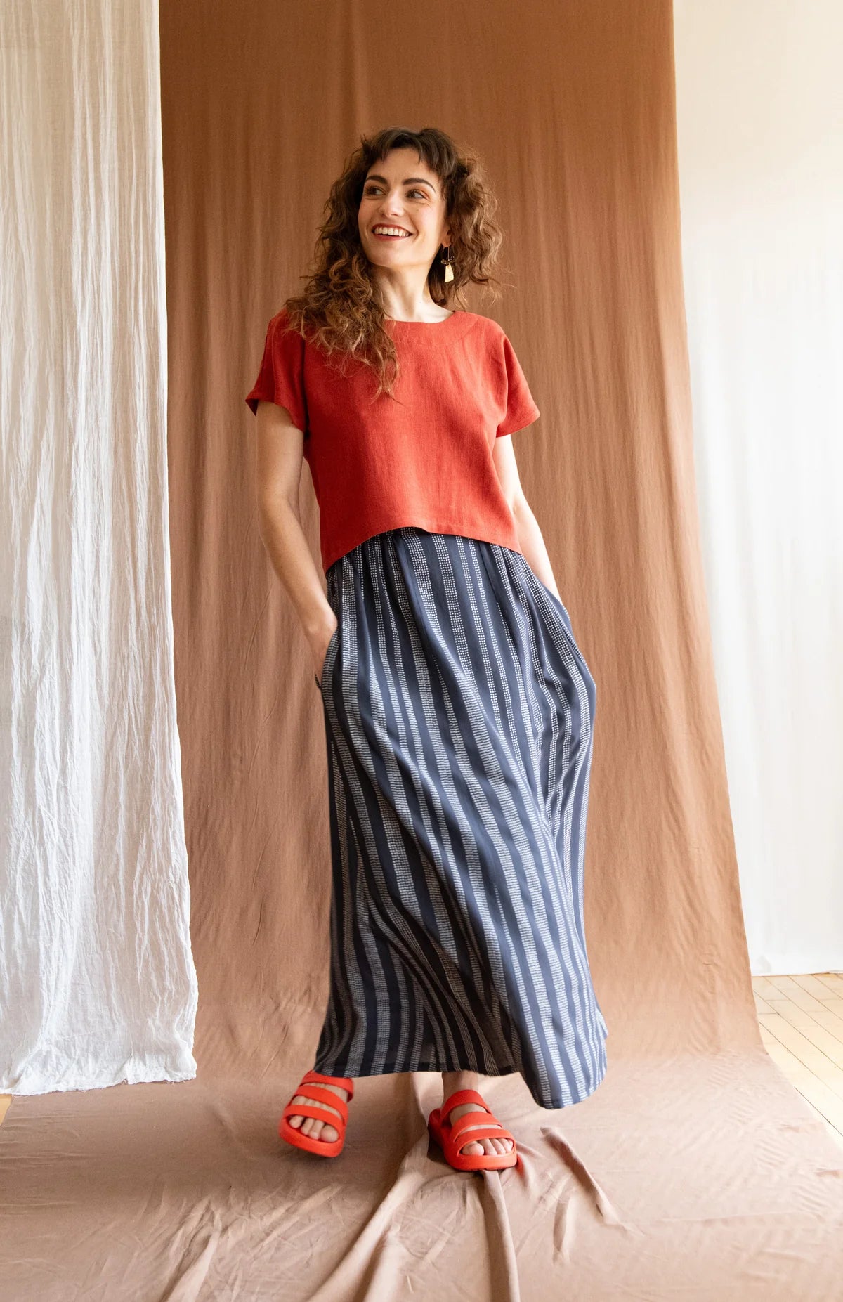 A woman wearing the Brendola Top by Kazak in Rust, a reversible top with a v-neck and snap front on one side and a round neck on the other side. It has short extended sleeves and a relaxed, cropped fit. She is wearing it with the Verona Skirt in Charcoal Dots and standing in front of a brown curtain.