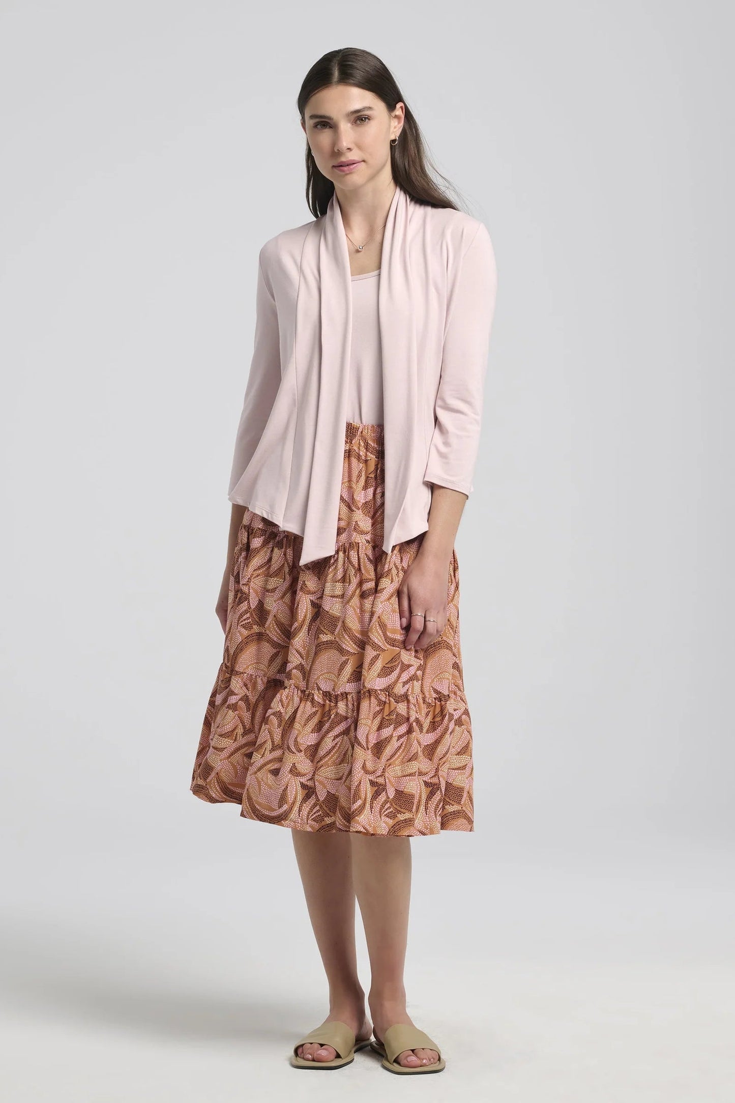 A woman wearing the Al-Uzza Skirt by Kollontai in Cinnamon print, a tiered skirt with an elastic waist and a below-the-knee length, with a pink top, standing in front of a white background