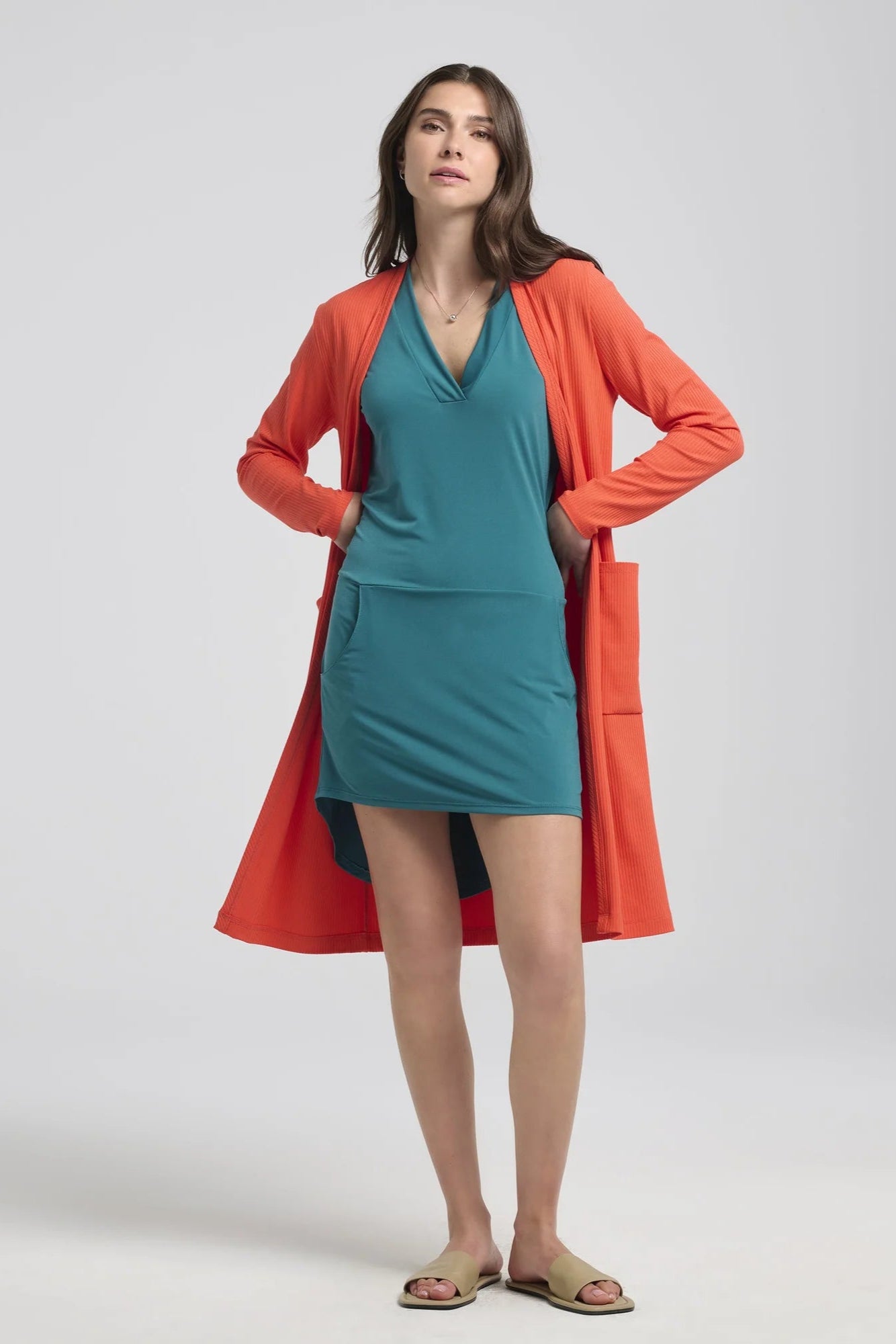 A woman wearing the Morticia Dress by Kollontai in Ocean, a sleeveless dress with a V-neck, a hi-low hemline, and a front pouch pocket. She is wearing it with the Strelisksi Vest in Papaya and standing in front of a white background.