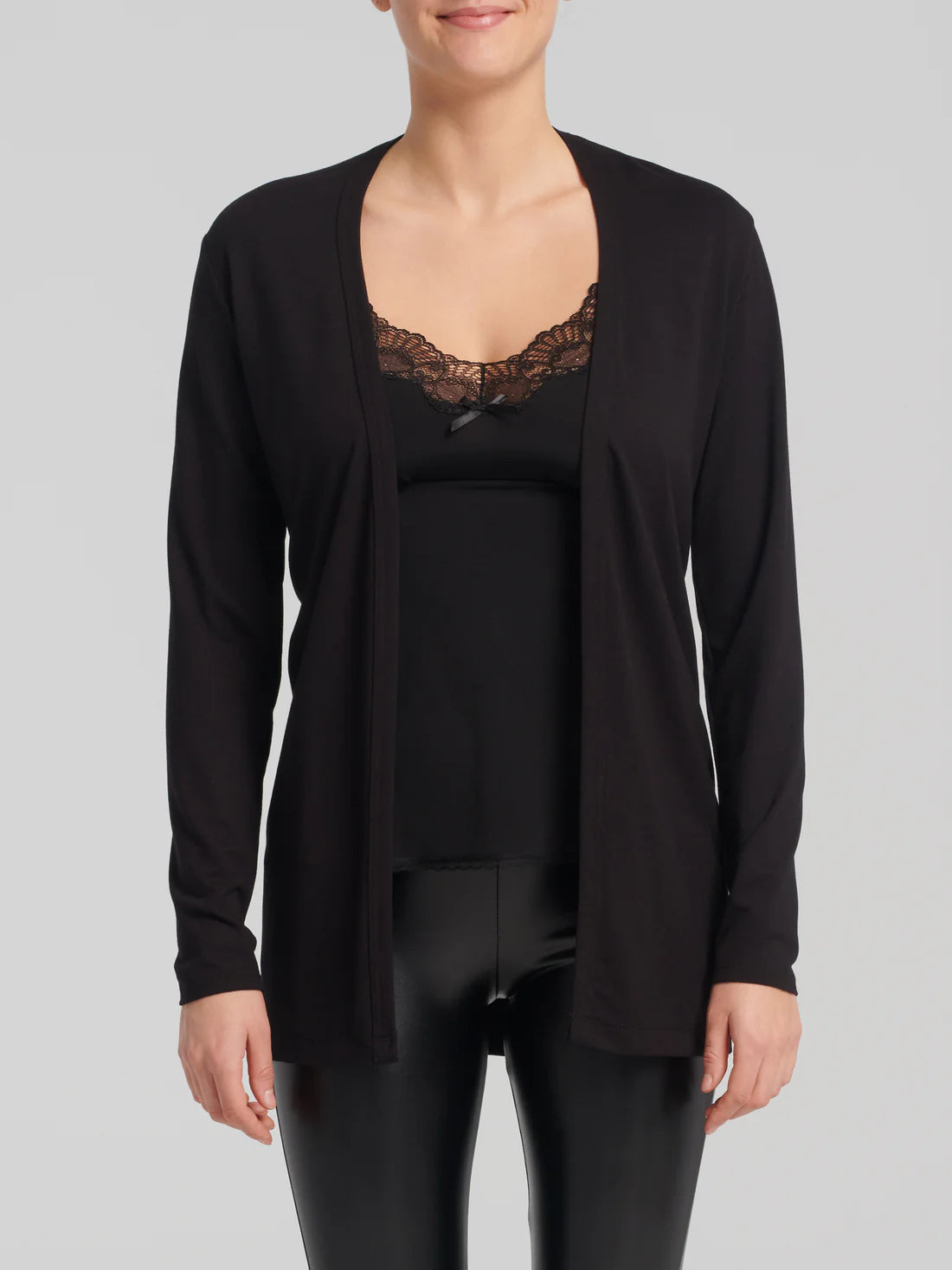A woman wearing the Umay Cardigan by Kollontai in Black, an open hip-length cardigan made from light viscose. She is wearing it over a black tank and black leggings and is standing in front of a white background.