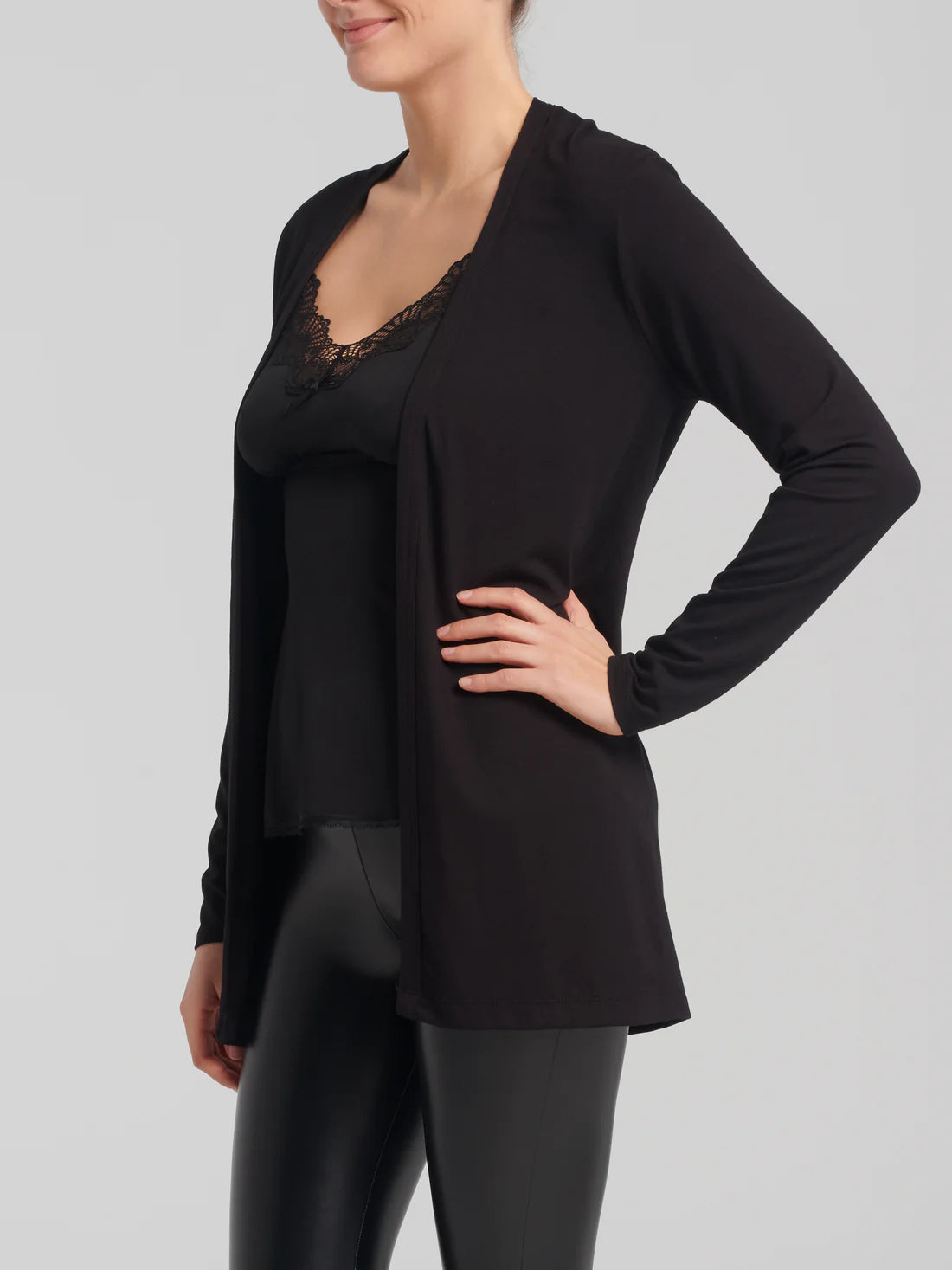 Side view of a woman wearing the Umay Cardigan by Kollontai in Black, an open hip-length cardigan made from light viscose. She is wearing it over a black tank and black leggings and is standing in front of a white background.