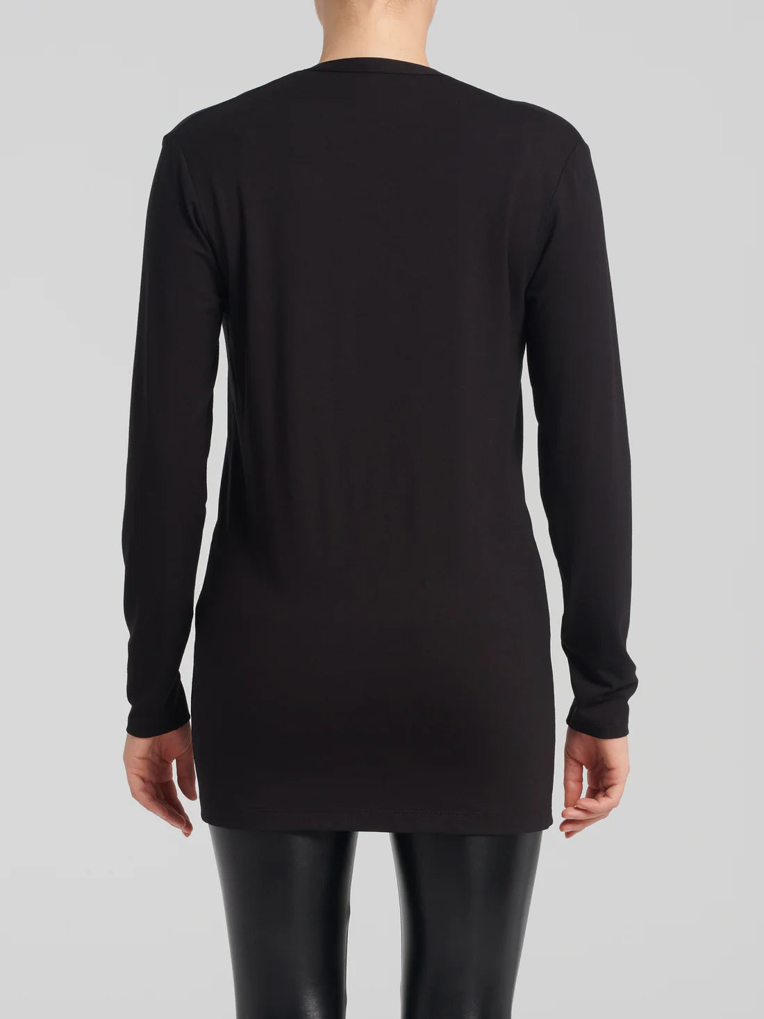 Back view of a woman wearing the Umay Cardigan by Kollontai in Black, an open hip-length cardigan made from light viscose. She is wearing it over a black tank and black leggings and is standing in front of a white background.