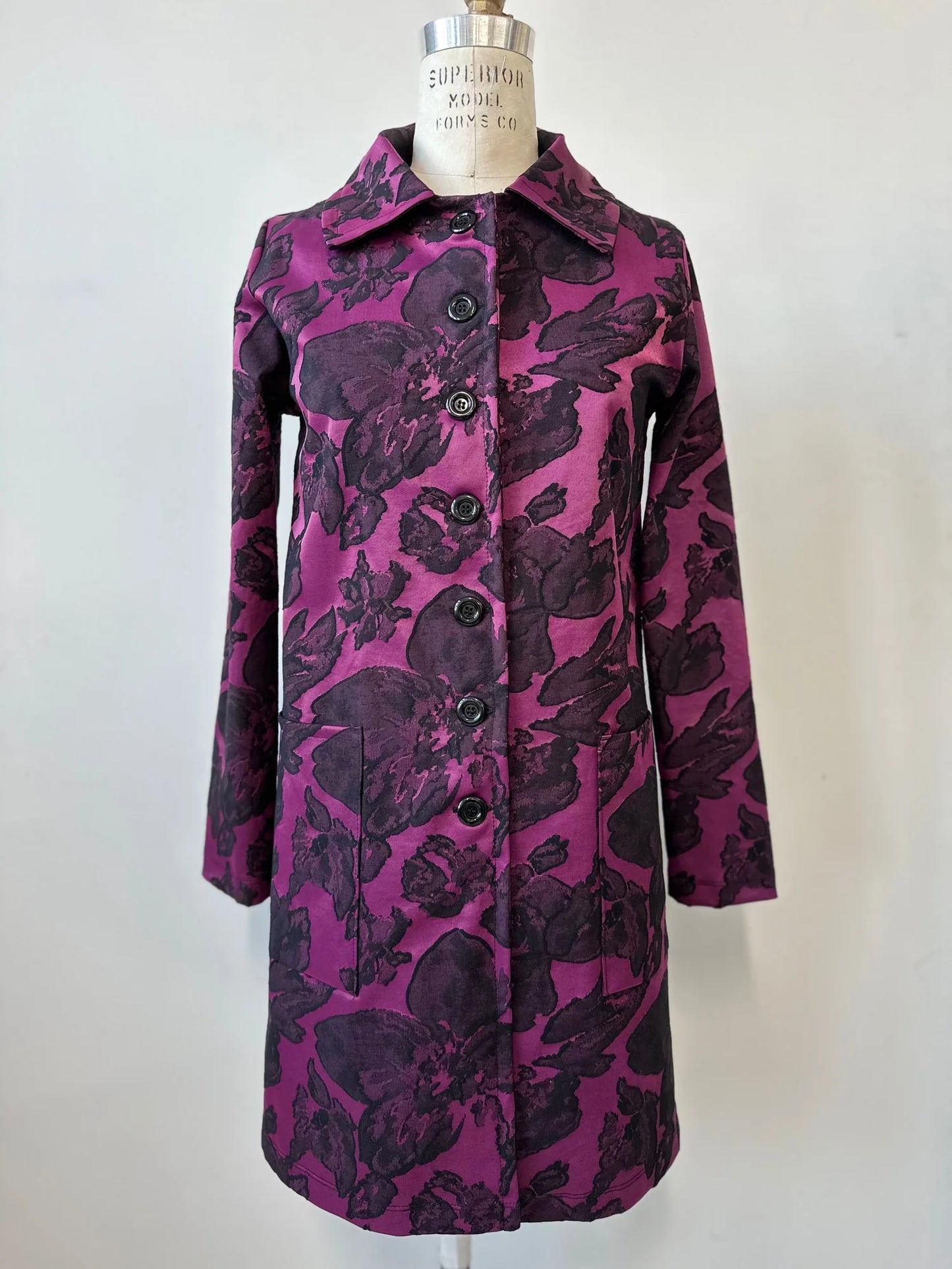 The Amarante Coat by Kollontai in Blackberry is shown on a mannequin against a white background. It's a satin coat with an intricate floral pattern, a classic collar, button front, straight cut that hits above the knee, and large pockets.