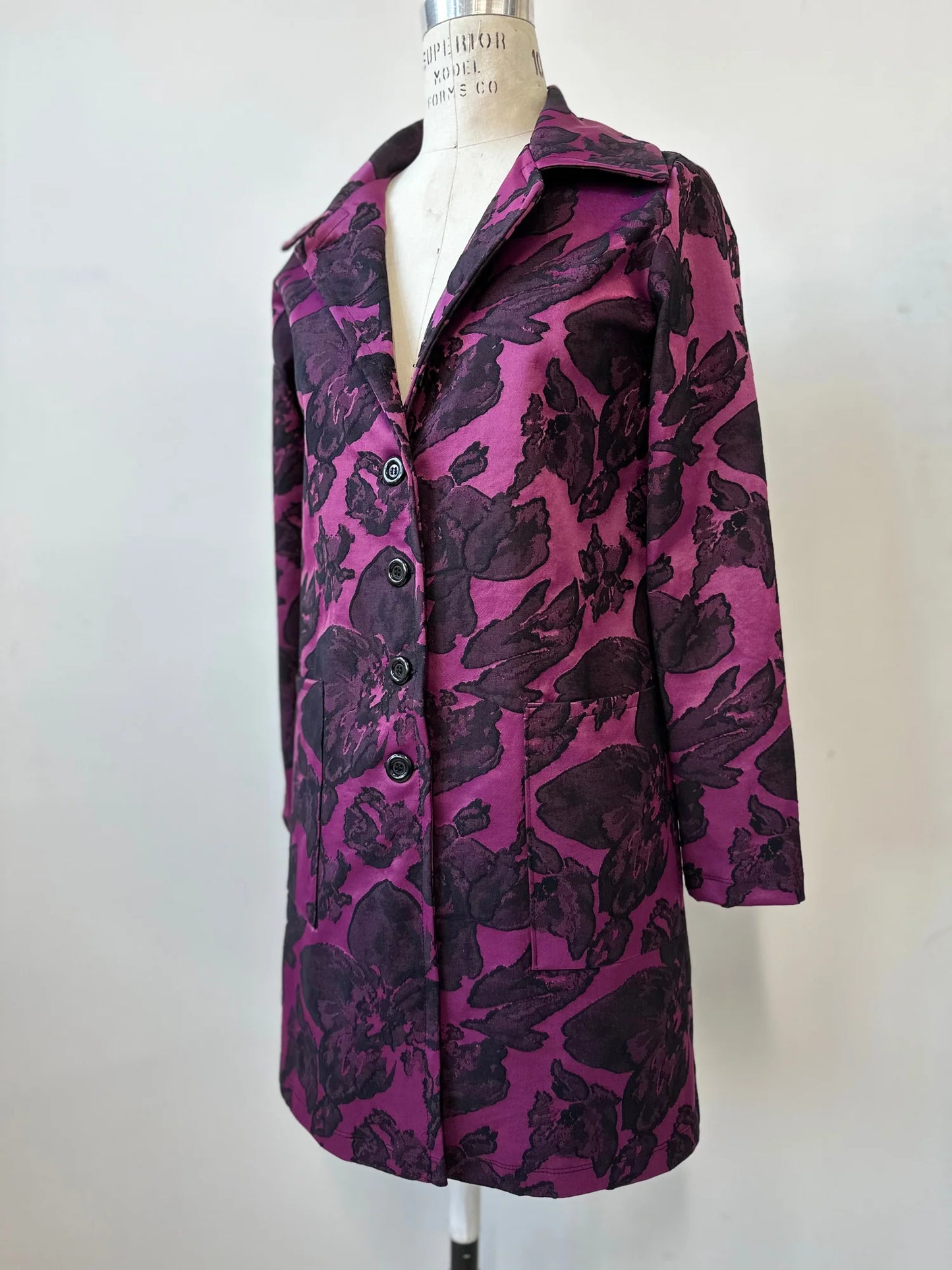 The Amarante Coat by Kollontai in Blackberry is shown on a mannequin against a white background. It's a satin coat with an intricate floral pattern, a classic collar, button front, straight cut that hits above the knee, and large pockets.