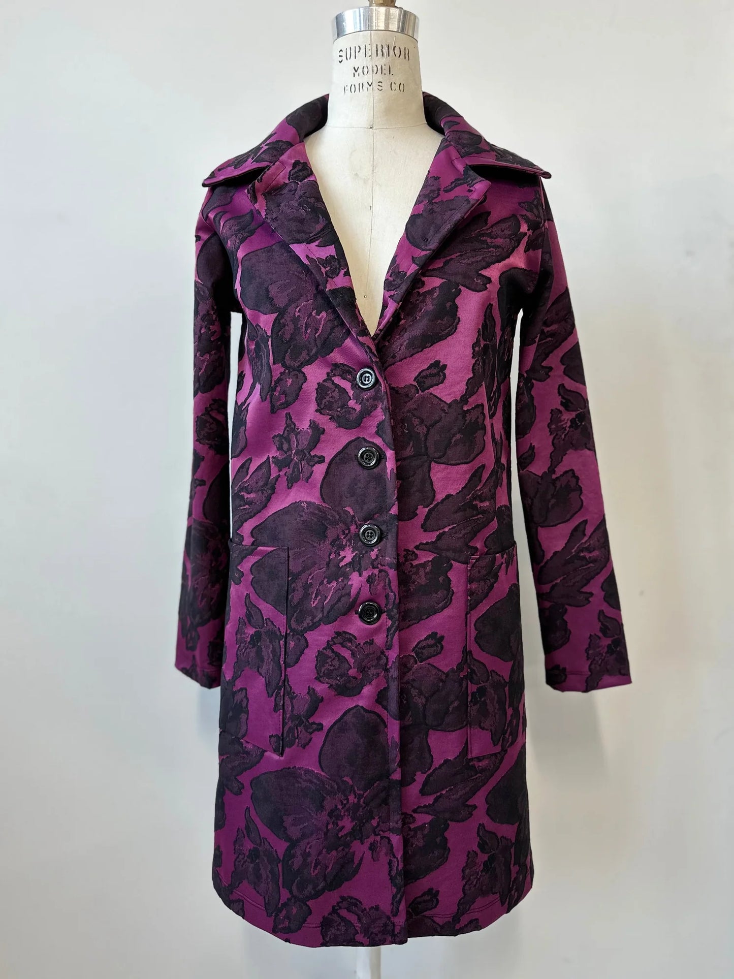 The Amarante Coat by Kollontai in Blackberry is shown on a mannequin against a white background. It's a satin coat with an intricate floral pattern, a classic collar, button front, straight cut that hits above the knee, and large pockets.