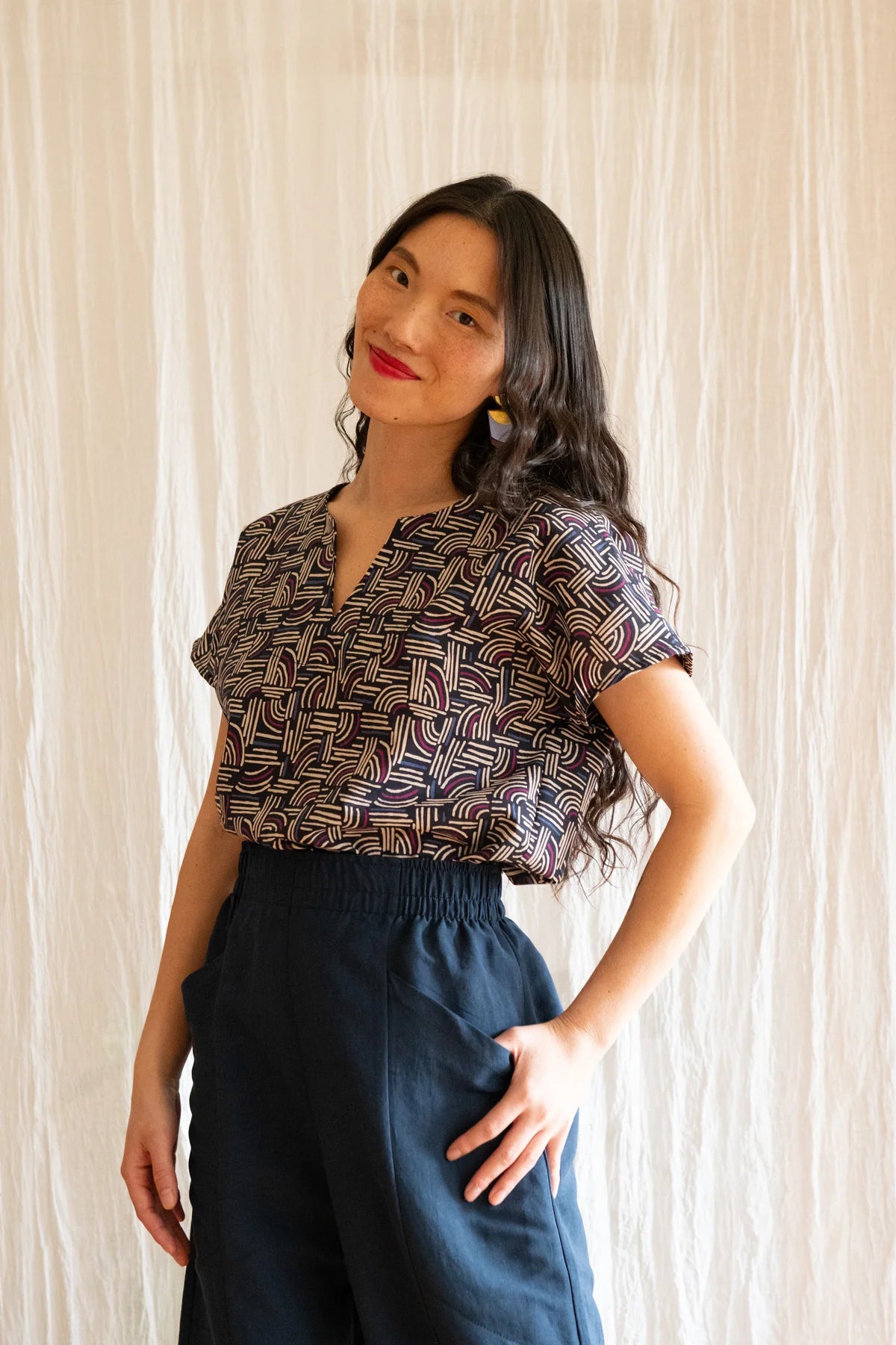 3/4 view of A woman wearing the Padua Pants by Kazak in Indigo, with a high waist, flat waistband at the front and elastic at the bag, wide legs, and large slanted pockets. She is wearing them with the Lucca Top in Night Basketwork and standing in front of a white curtain.