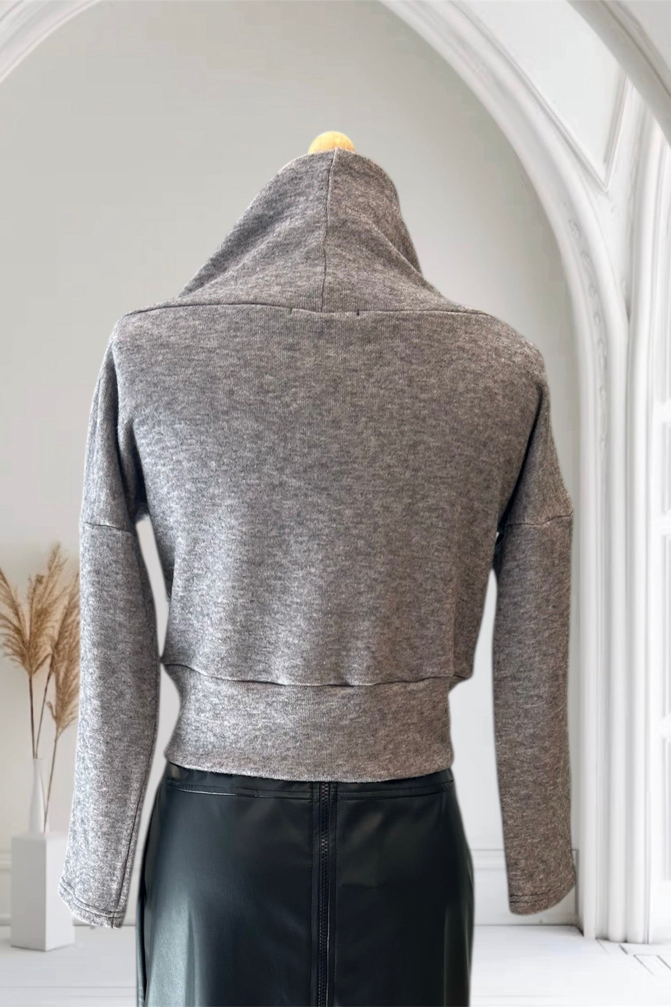 A back view of the changes Sweater by Allison Wonderland in Light Grey, featuring a large cowl neck, long dropped sleeves, and a wide waistband, is shown with the Desire Skirt in Black against a white arched backdrop.