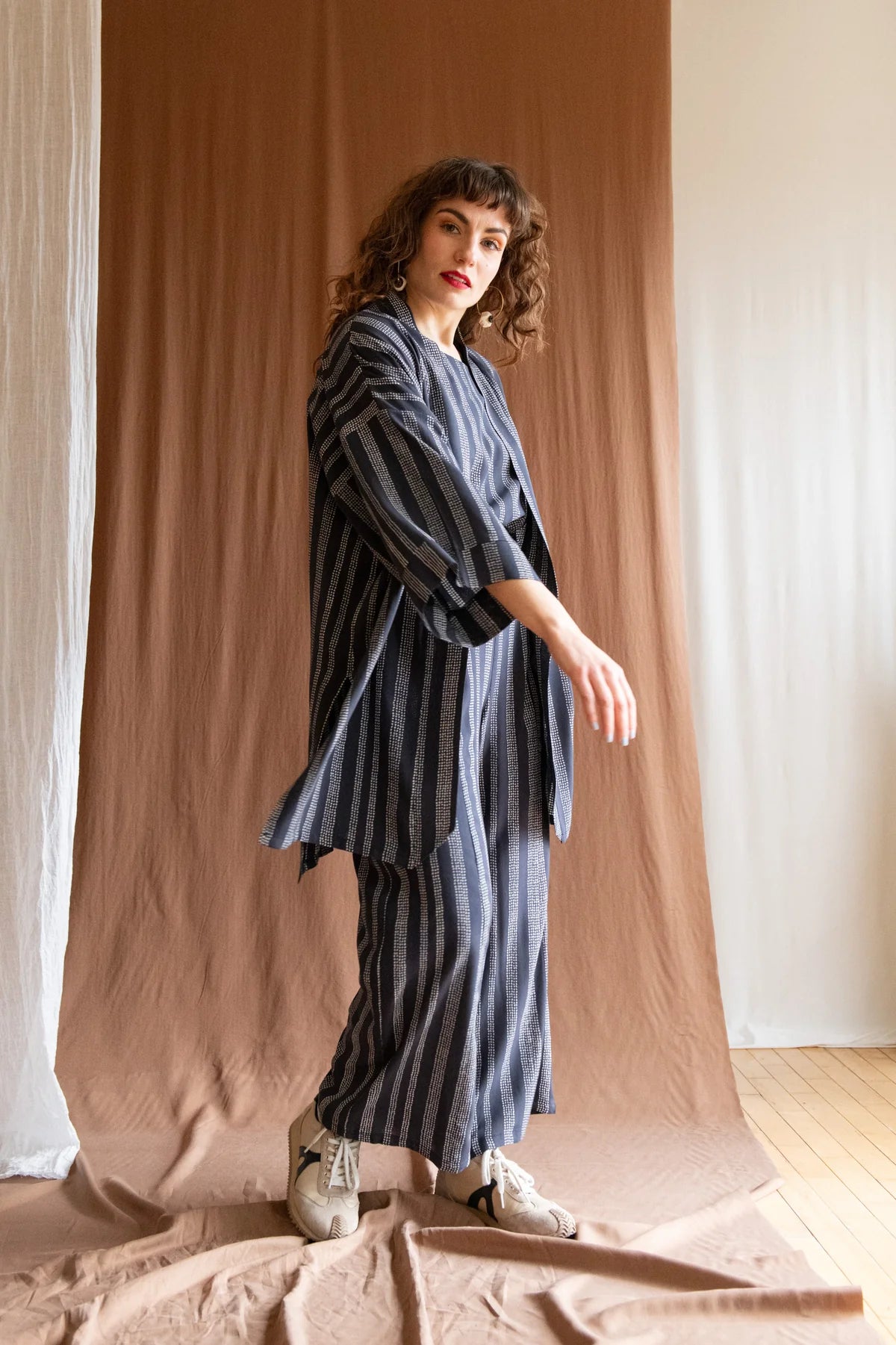 A woman wearing the Padua Pants by Kazak in Charcoal Dots, with a high waist, flat waistband at the front and elastic at the bag, wide legs, and large slanted pockets. She is wearing them with the matching Sandrigo top and Mirano Jacket and standing in front of a white curtain.