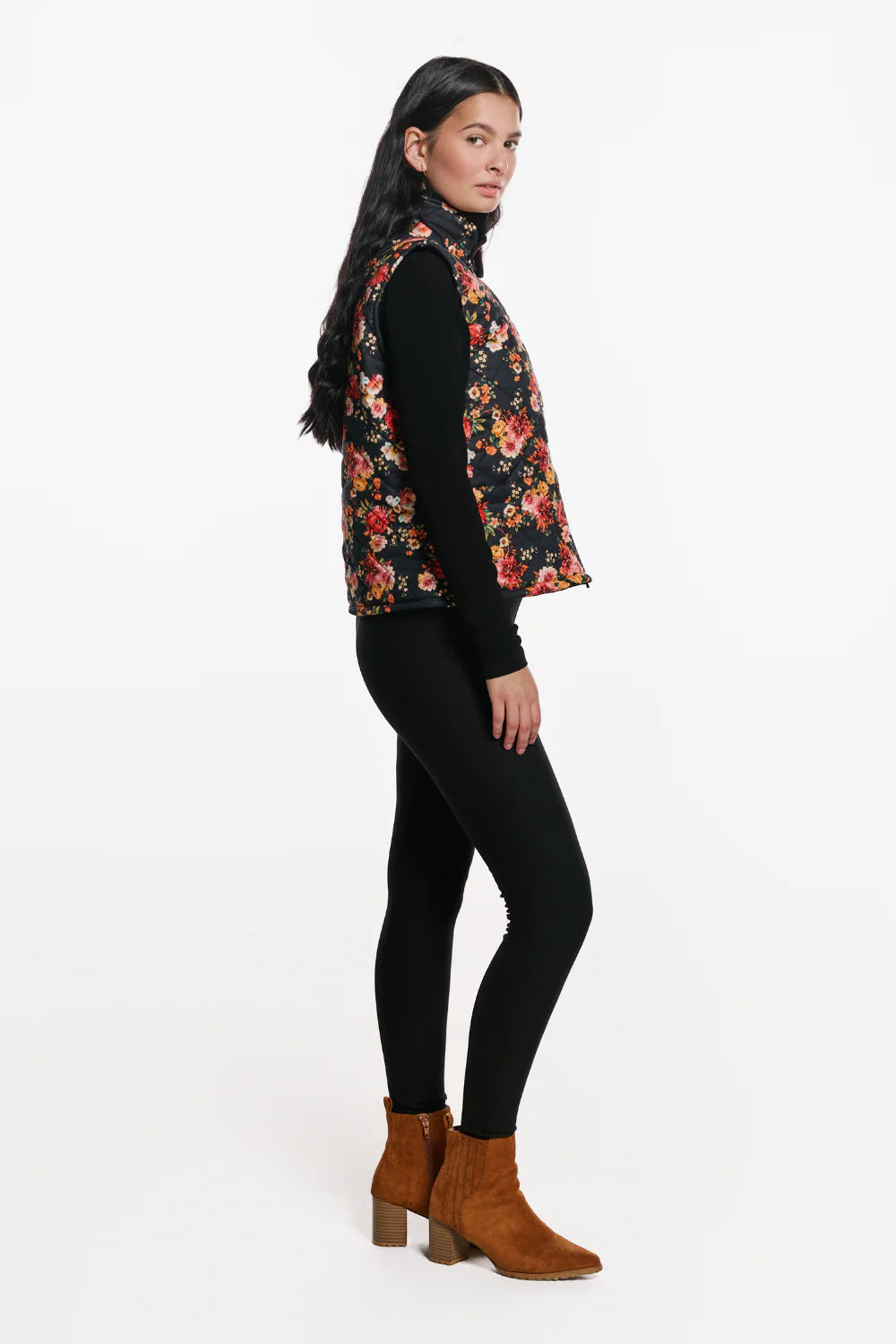 Side view of a woman wearing the Harper Vest by EMK, a quilted floral vest with a zip front two pockets, and black waffle knit fabric at the collar. She is wearing it with black pants and standing in front of a white background.