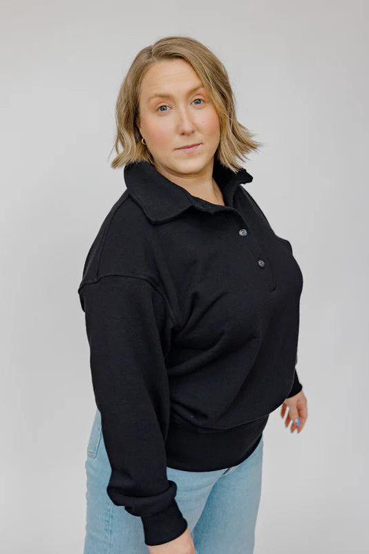 A woman wearing the Classic Coastal Sweater by Blondie in Black, a relaxed fit sweatshirt with a collar and four button placket. She is wearing it with jeans and standing in front of a white background.