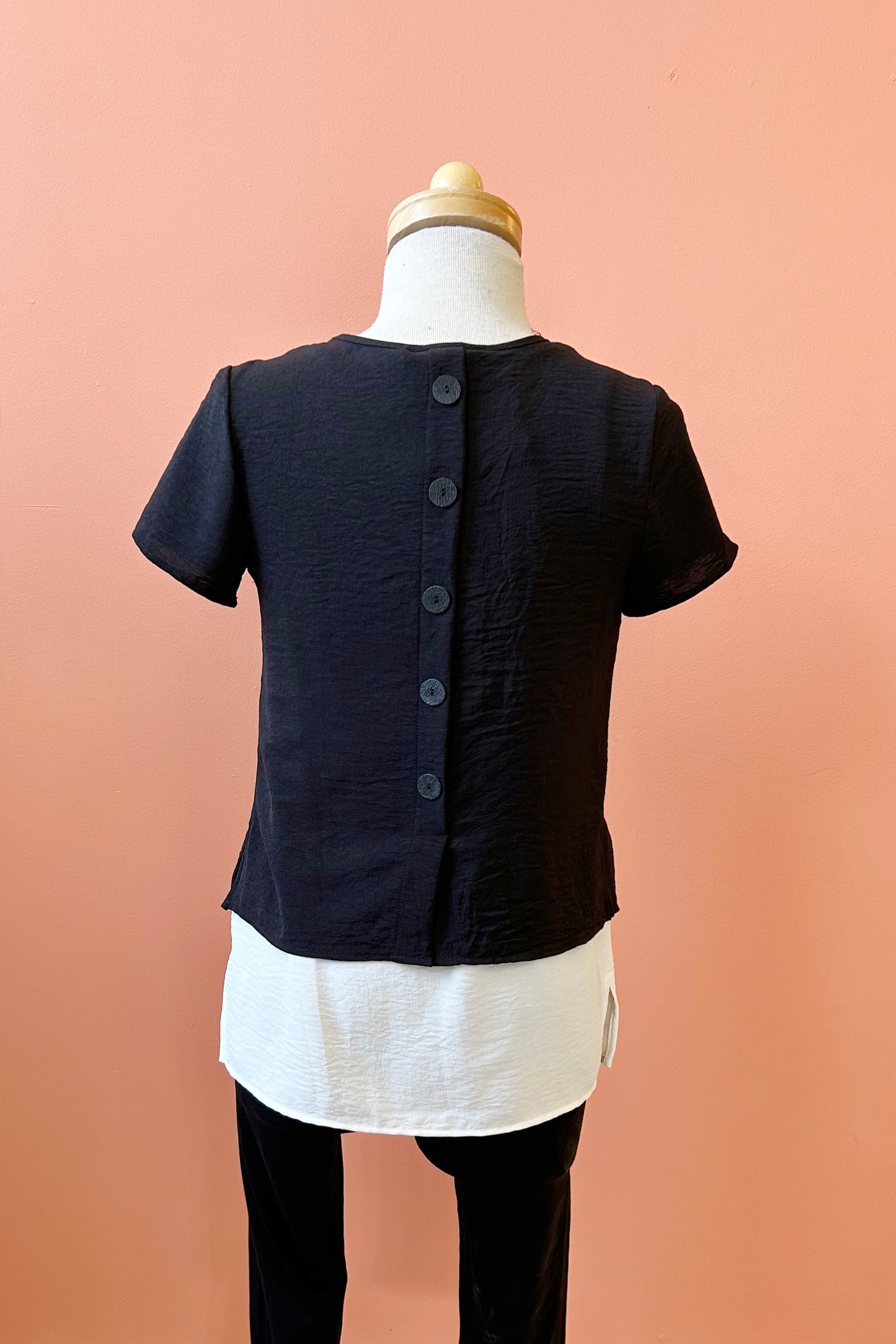 A back view of the Oona Top by Compli K in Black/Ivory, featuring an over-shirt with buttons up the back and a patch pocket with a button over a longer tank, is shown on a mannequin against a coral wall.