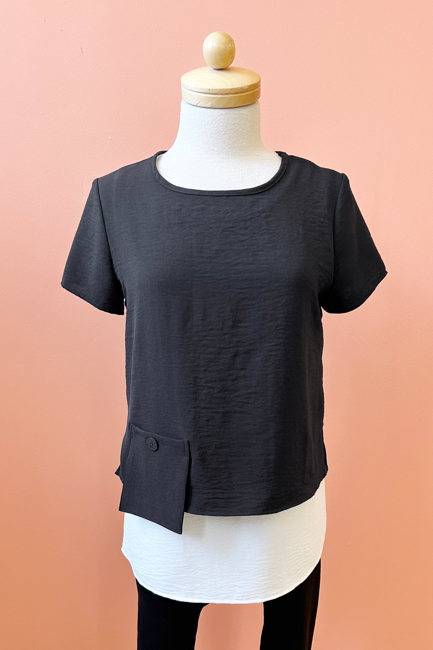 The Oona Top by Compli K in Black/Ivory, featuring a over-shirt with buttons up the back and a patch pocket with a button over a longer tank, is shown on a mannequin against a coral wall.