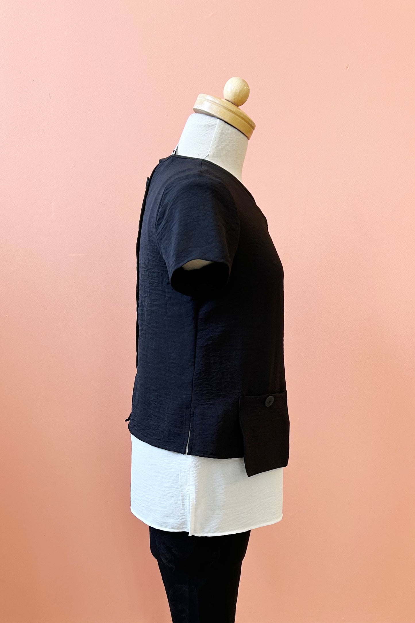 A side view of the Oona Top by Compli K in Black/Ivory, featuring an over-shirt with buttons up the back and a patch pocket with a button over a longer tank, is shown on a mannequin against a coral wall.