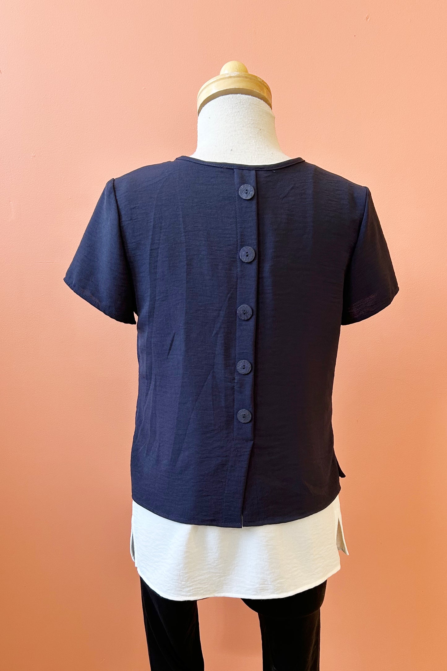 A back view of the Oona Top by Compli K in Navy/Ivory, featuring an over-shirt with buttons up the back and a patch pocket with a button over a longer tank, is shown on a mannequin against a coral wall.