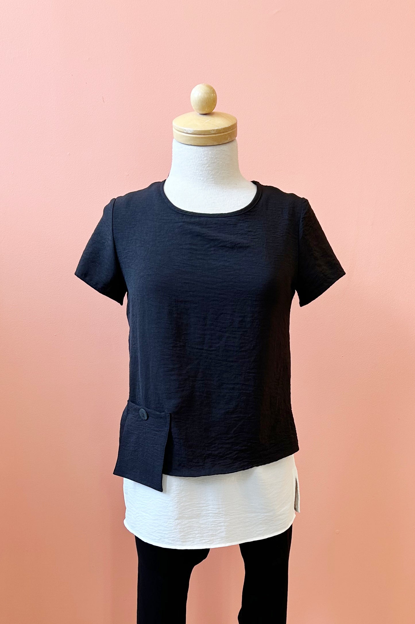 The Oona Top by Compli K in Navy/Ivory, featuring an over-shirt with buttons up the back and a patch pocket with a button over a longer tank, is shown on a mannequin against a coral wall.