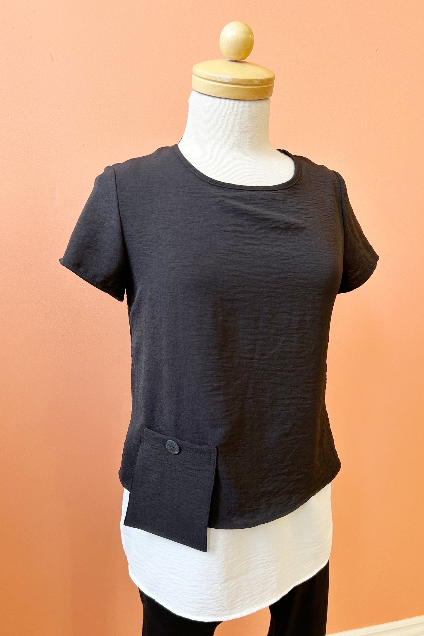 The Oona Top by Compli K in Black/Ivory, featuring an over-shirt with buttons up the back and a patch pocket with a button over a longer tank, is shown on a mannequin against a coral wall.
