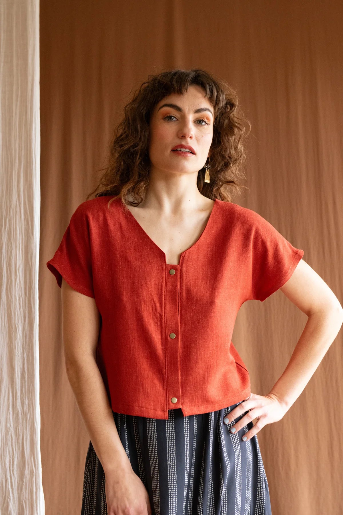 3/4 view of a woman wearing the Brendola Top by Kazak in Rust, a reversible top with a v-neck and snap front on one side and a round neck on the other side. It has short extended sleeves and a relaxed, cropped fit. She is wearing it with the Verona Skirt in Charcoal Dots and a patterned skirt and standing in front of a brown curtain.