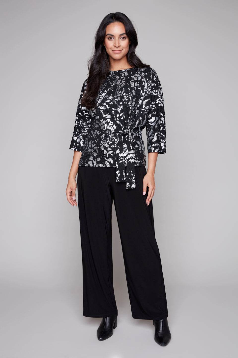 A woman wearing the Posey Sweater from Compli K in Black and Silver, with a round neck, 3/4 sleeves, and a tie detail at the waist, with black pants. She is standing in front of a grey background.