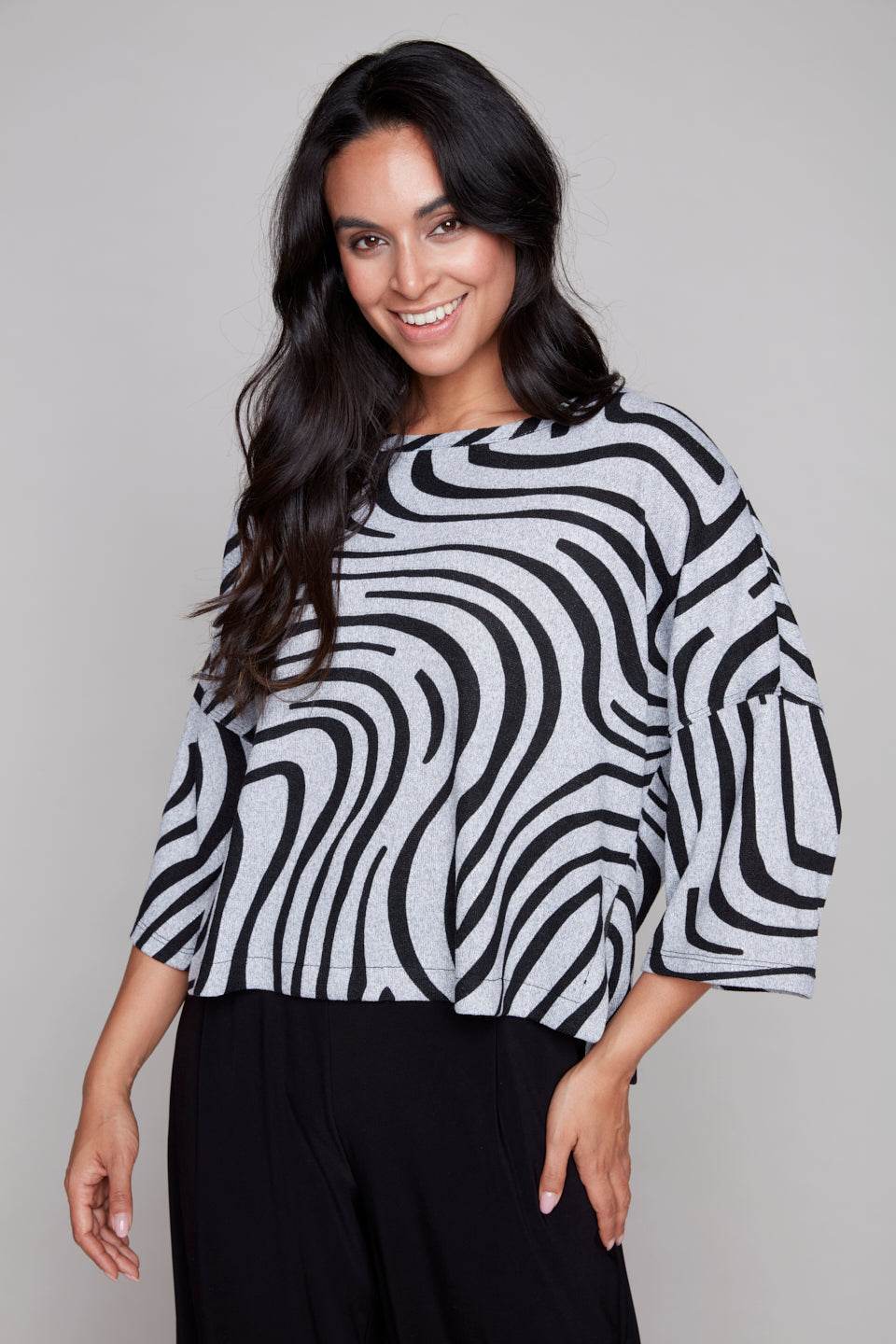 A woman wearing the Paula Top by Compli K in Silver Black Swirl, with round neck, 3/4 drop sleeves, and a hip length, with black pants. She is standing in front of a grey background.