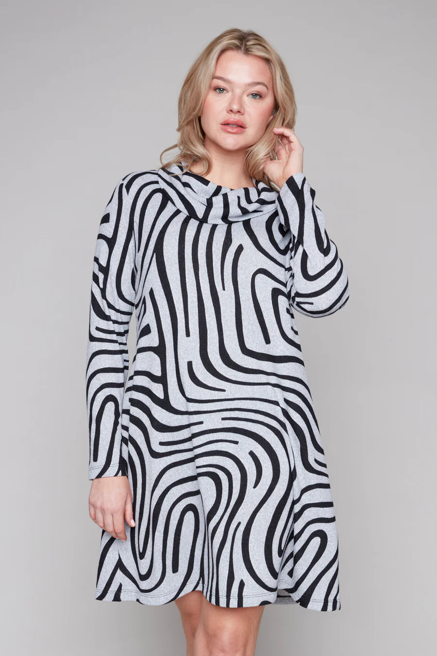 A woman wearing the Powell Dress by Compli K in Black and Silver Swirl, featuring a cowl neck, long sleeves, and a slight trapeze shape. She is standing in front of a grey background.