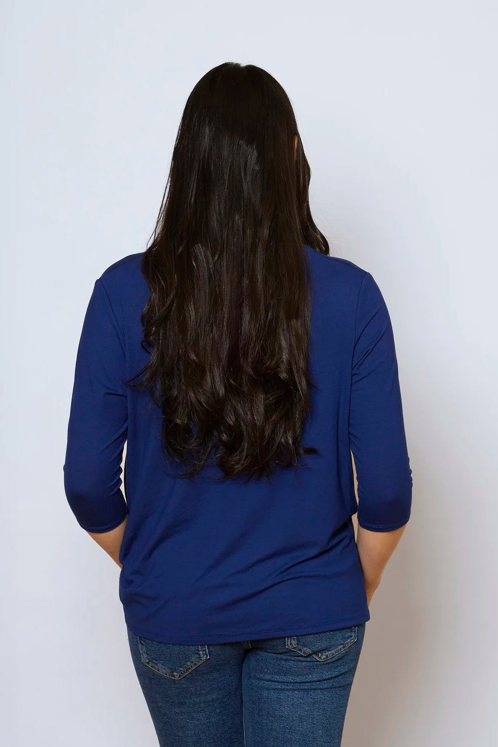 Back view of a woman wearing the Reese Top by Tangente in Blue, with a round neck, 3/4 drop sleeves, and a twisted hem in front. She is wearing it with jeans and standing in front of a white background.