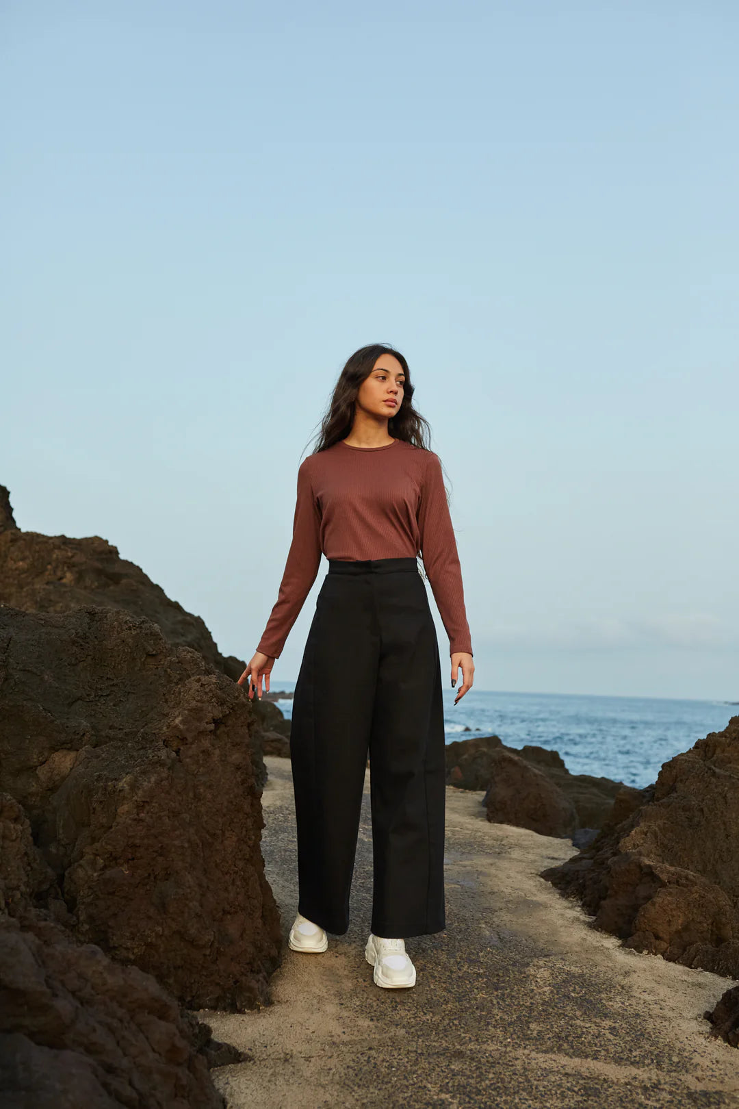 A woman wearing the Vienna Pants by MAS in Pepper, with a wide leg, side seams that sit slightly to the front, and two back pockets, with a rust coloured shirt. She is standing on a rocky path by the ocean.