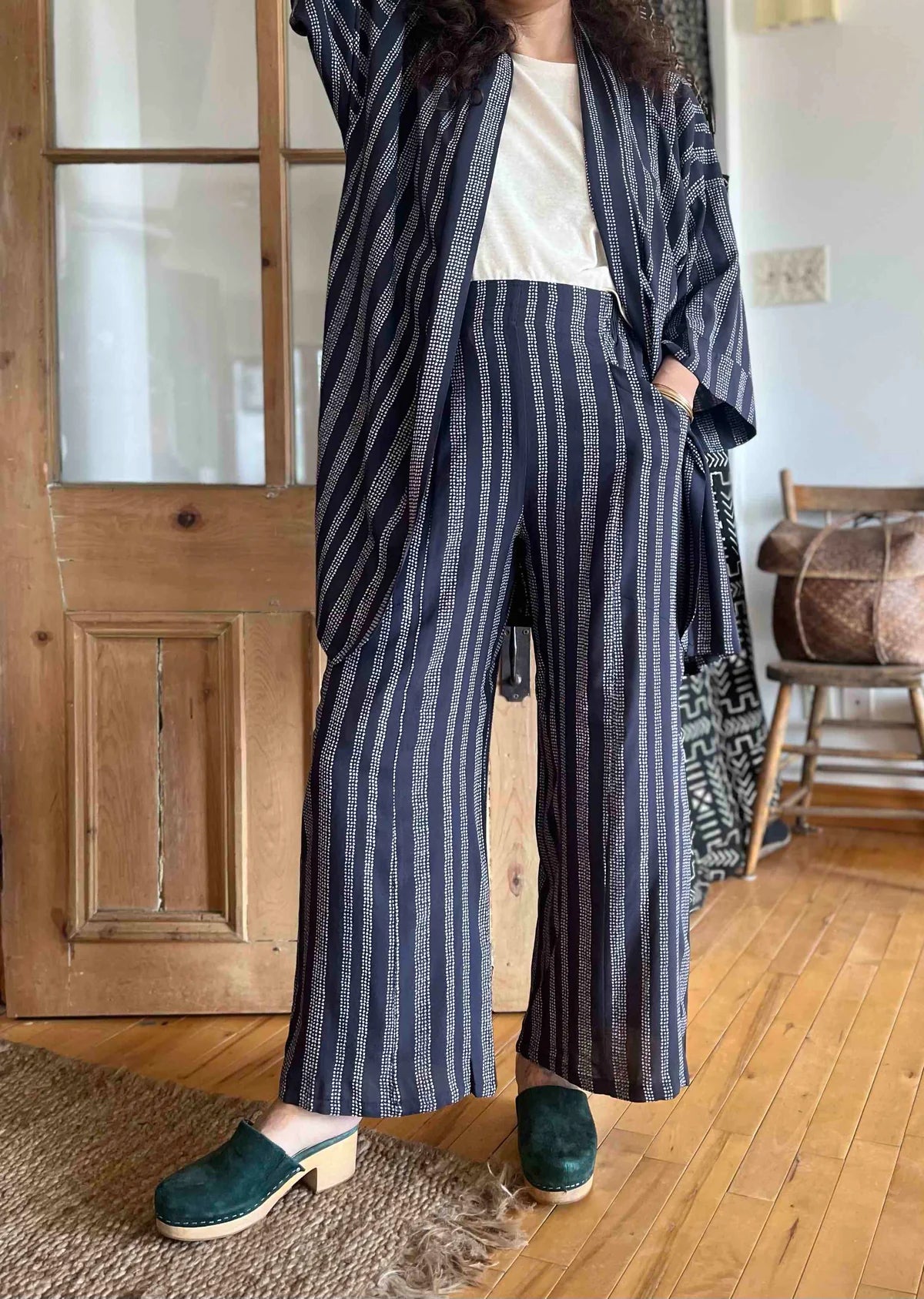 Neck down view of A woman wearing the Padua Pants by Kazak in Charcoal Dots, with a high waist, flat waistband at the front and elastic at the bag, wide legs, and large slanted pockets. She is wearing them with a white top and the matching Mirano Jacket and standing in front of a door.