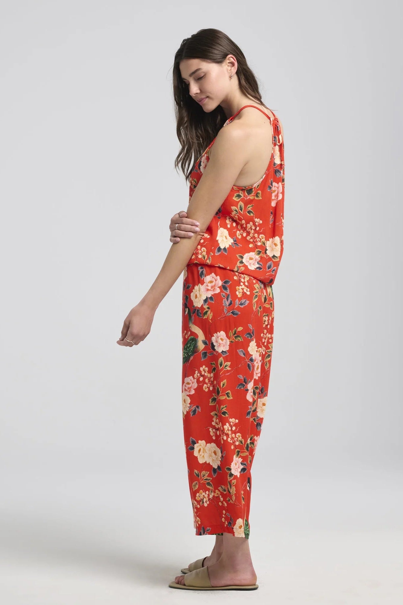 Side view of a woman wearing the Zephyrine Jumpsuit by Kollontain Red floral, with a halter neck, elastic waist with a bloused effect, and wide cropped legs. She is standing in front of a white background.