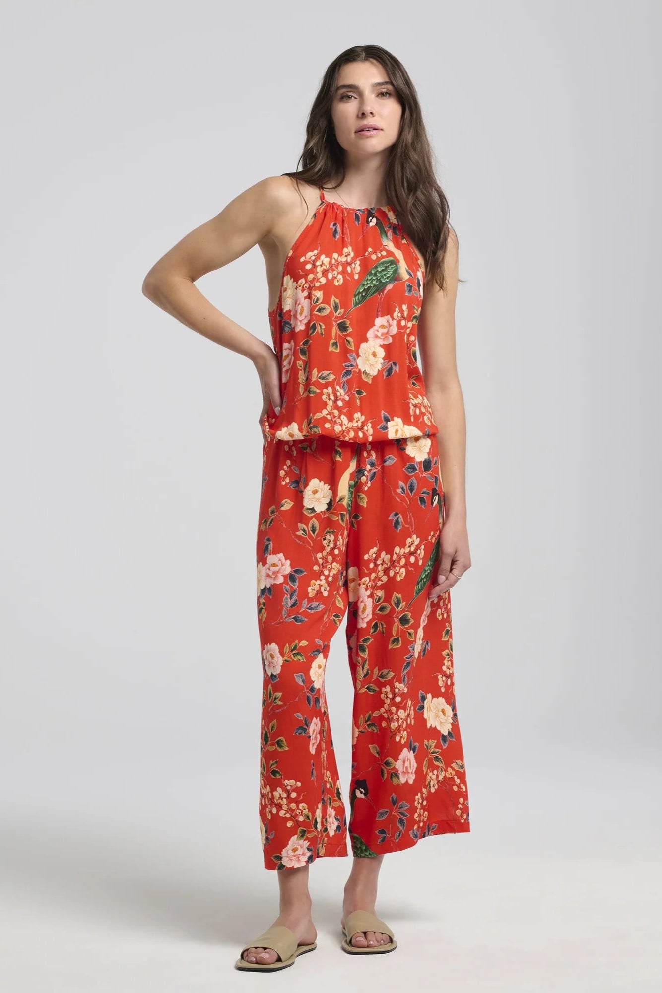 A woman wearing the Zephyrine Jumpsuit by Kollontain Red floral, with a halter neck, elastic waist with a bloused effect, and wide cropped legs. She is standing in front of a white background.