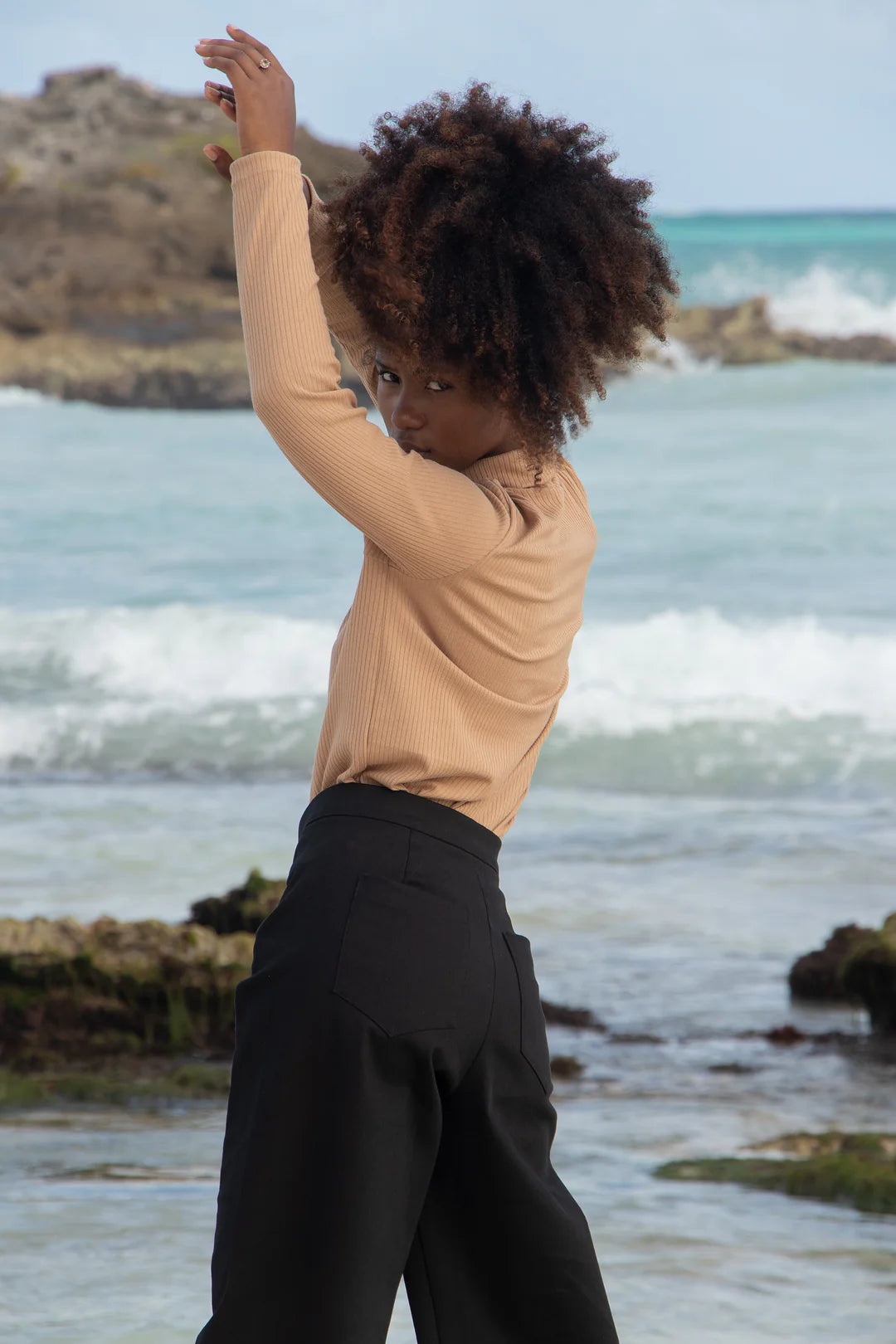 A woman wearing the Vienna Pants by MAS in Pepper with a wide leg, side seams that sit slightly to the front, and two back pockets, with tan shirt. She is standing in front of the ocean.